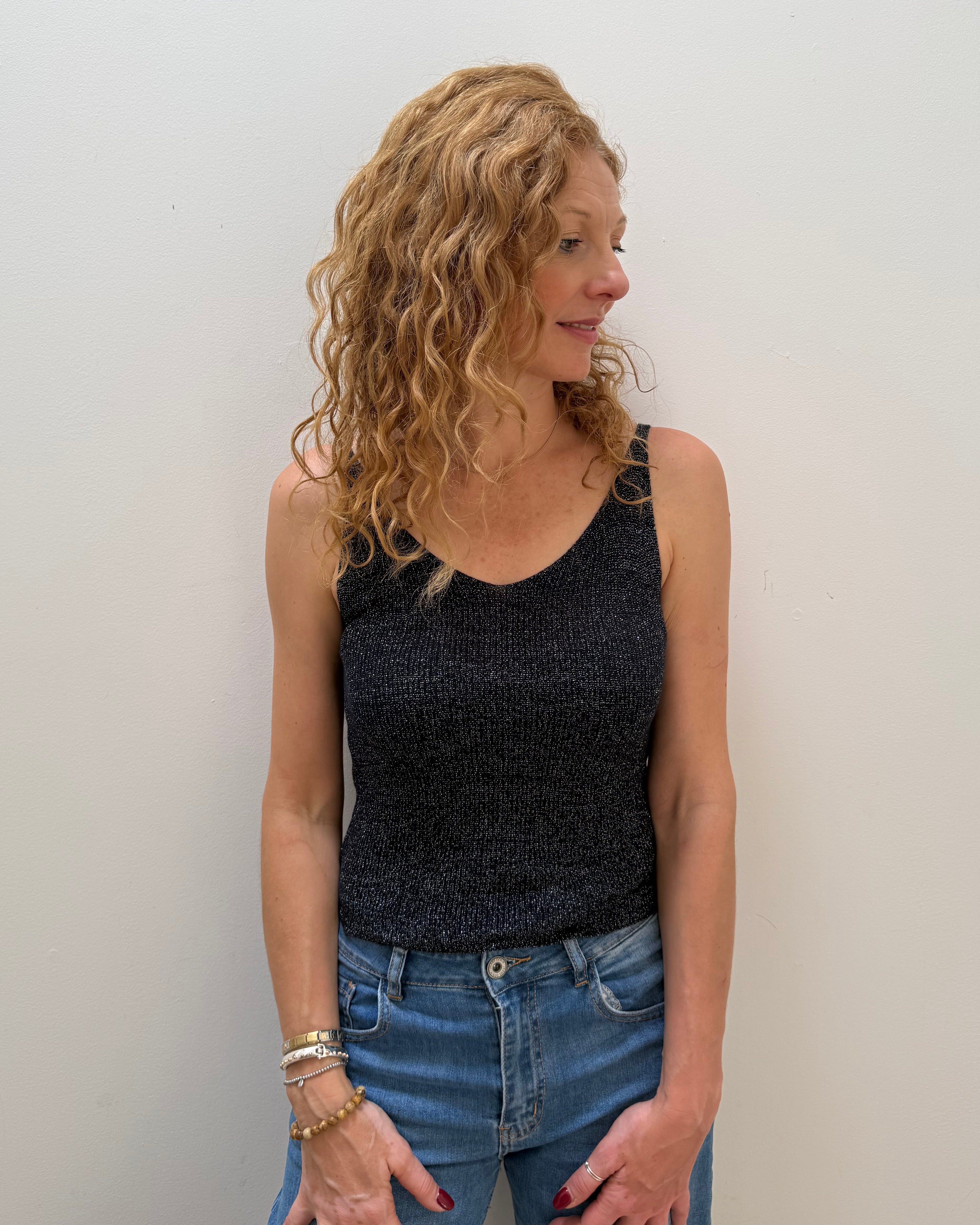 Woman with curly hair wearing a black sleeveless top and blue jeans against a white wall.
