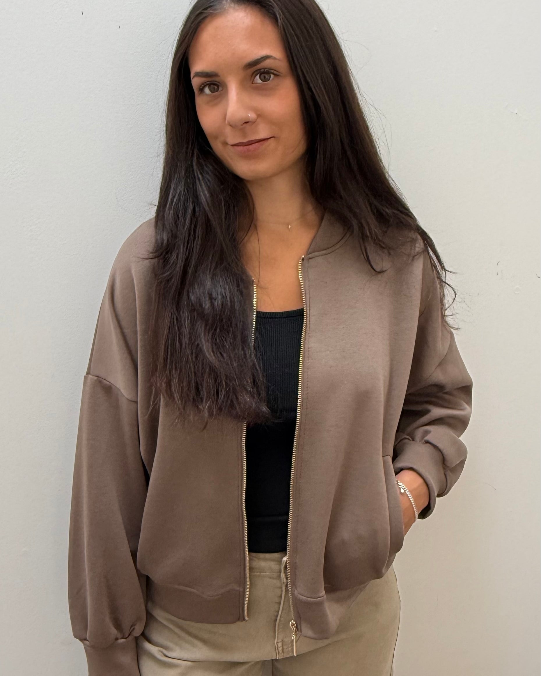 A woman with long dark hair wearing a taupe-brown bomber jacket with gold-tone zippers over a black top and beige pants, standing against a plain off-white wall.