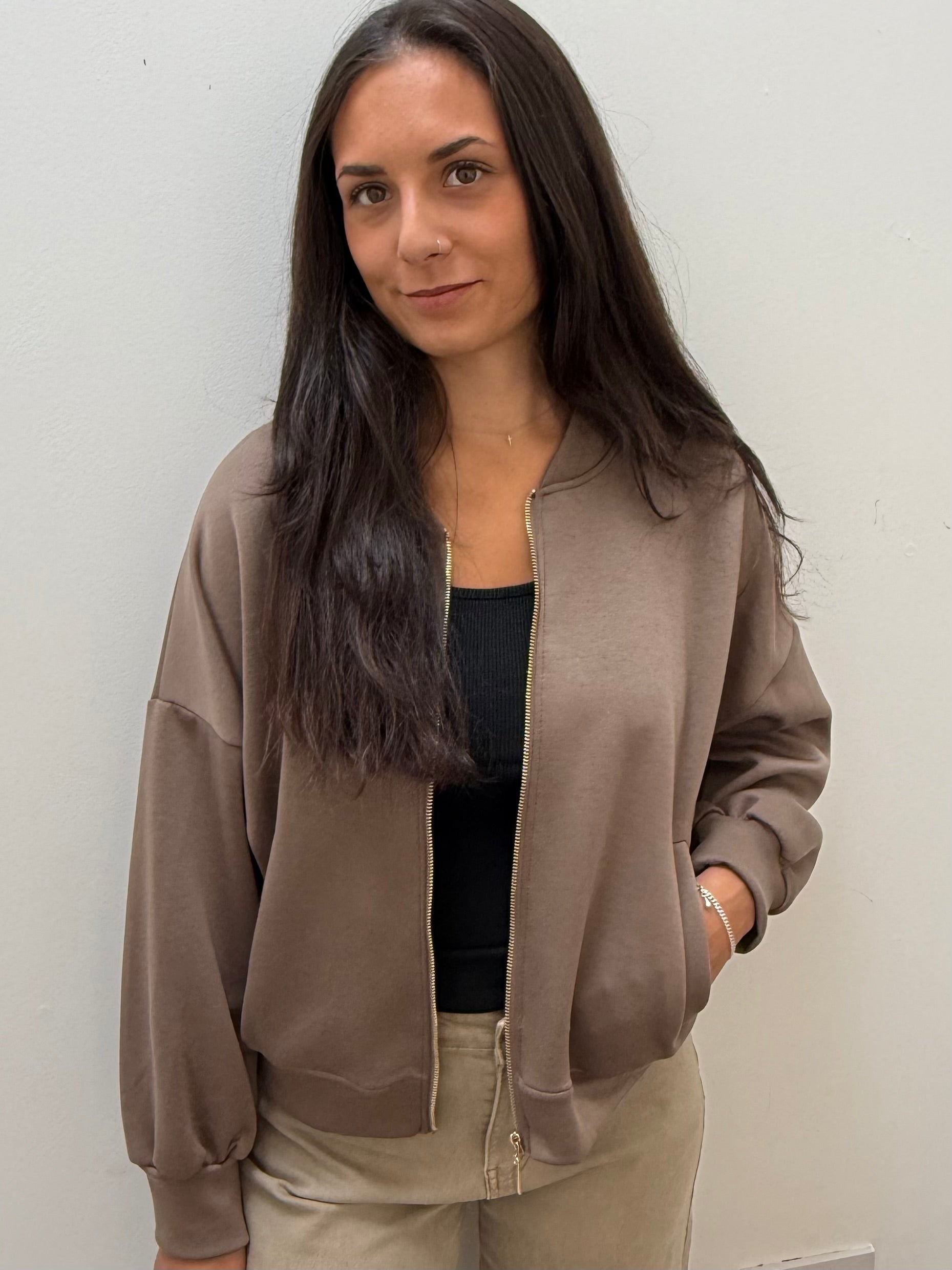 A woman with long dark hair wearing a taupe-brown bomber jacket with gold-tone zippers over a black top and beige pants, standing against a plain off-white wall.