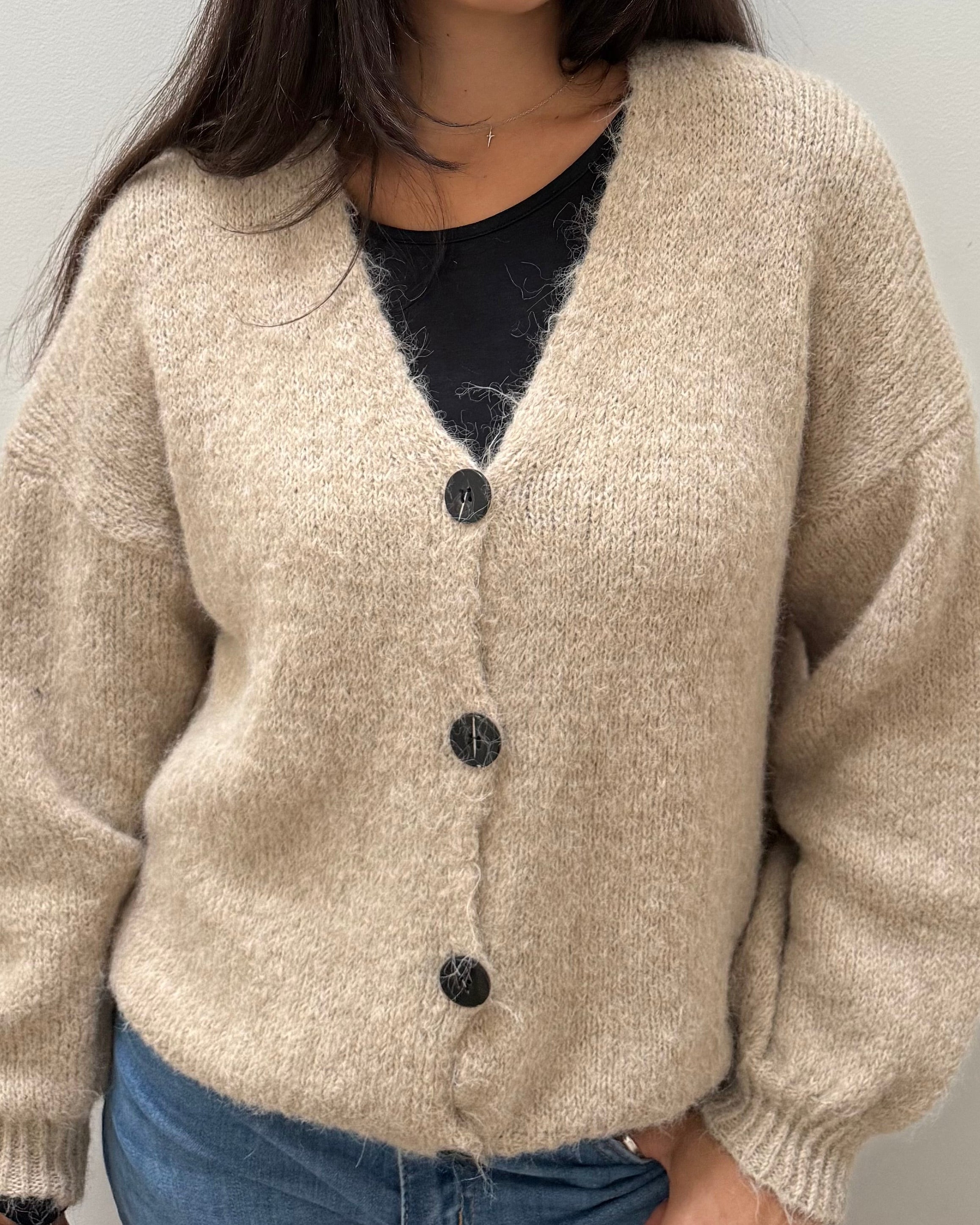 A woman wearing a beige, textured knit cardigan with dark buttons over a black top and blue jeans, standing against a plain light gray wall.