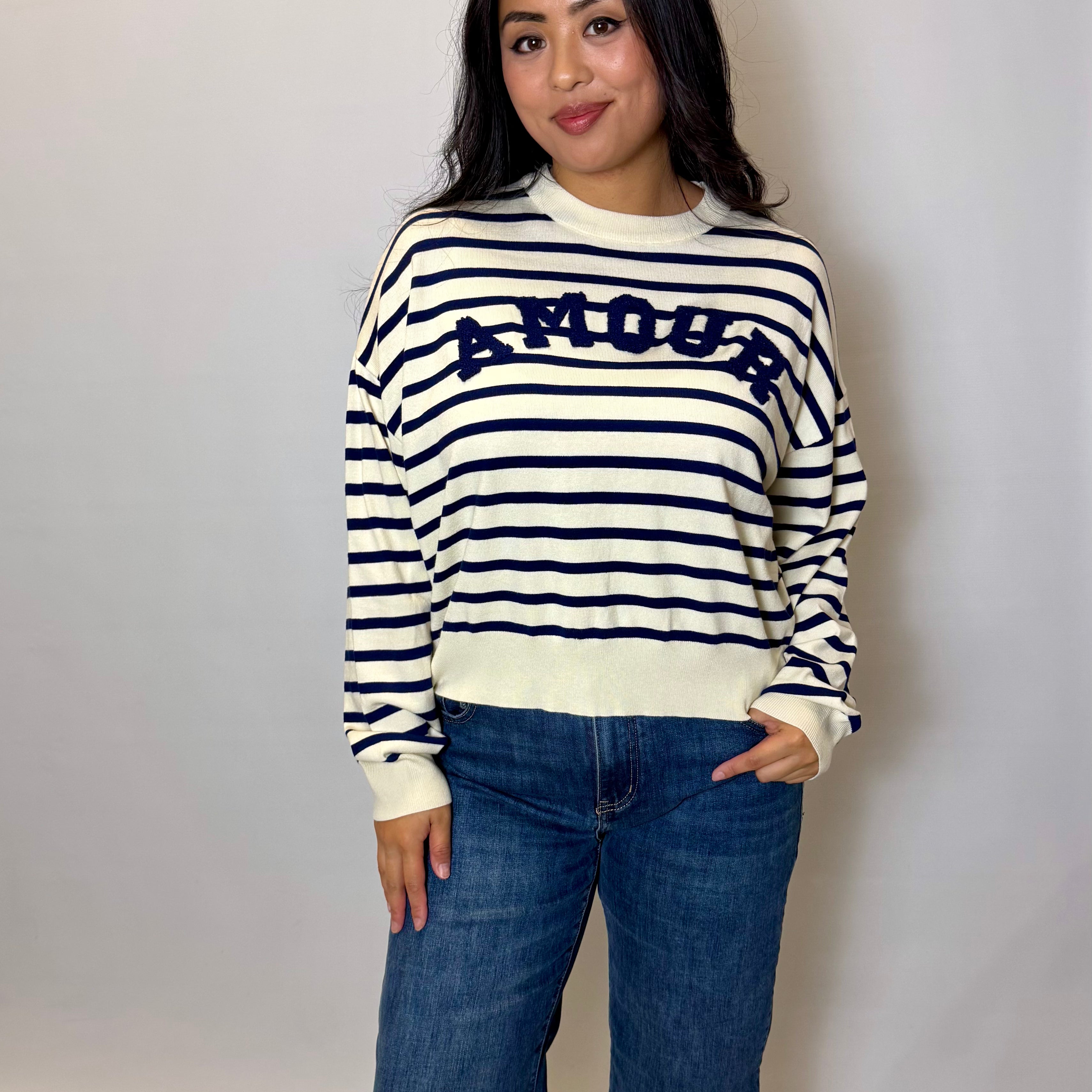 Woman wearing a striped sweater with 'AMOUR' text and jeans against a plain background