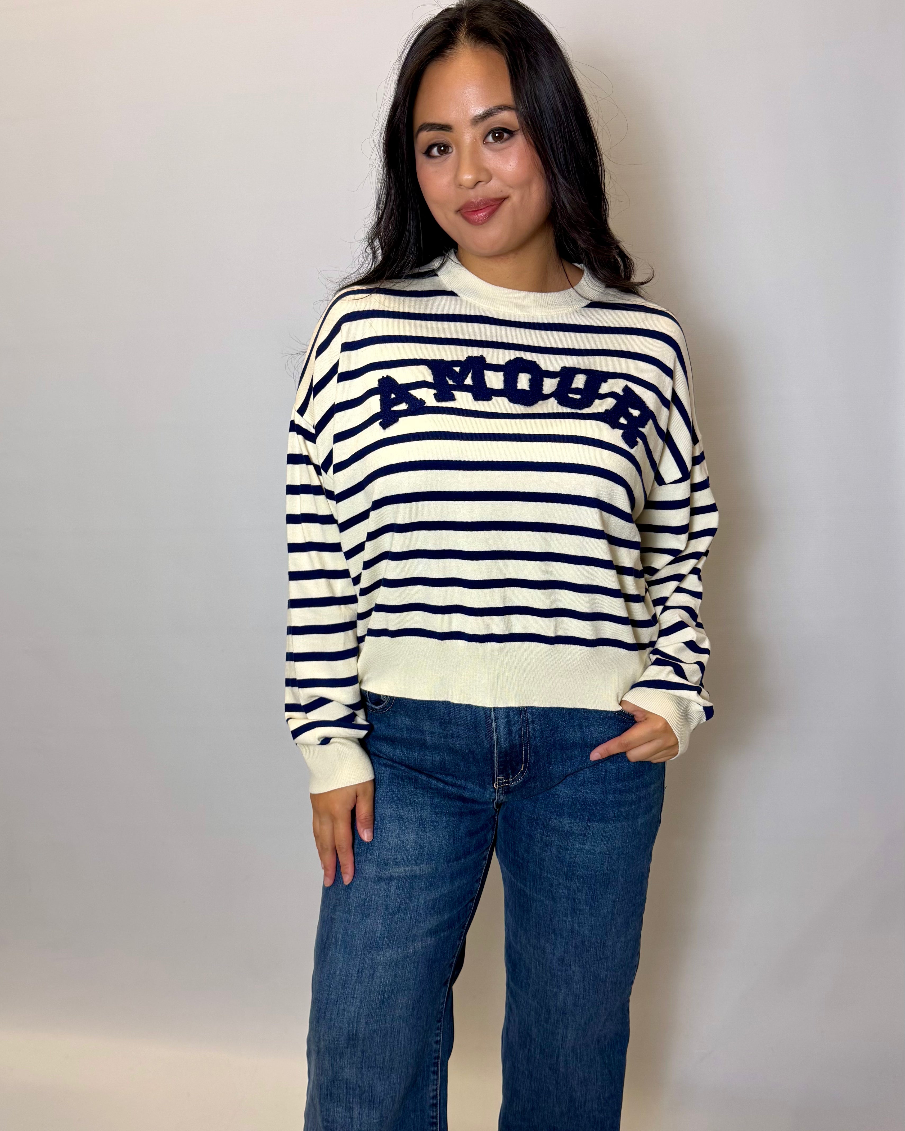 Woman wearing a striped sweater with 'AMOUR' text and jeans against a plain background