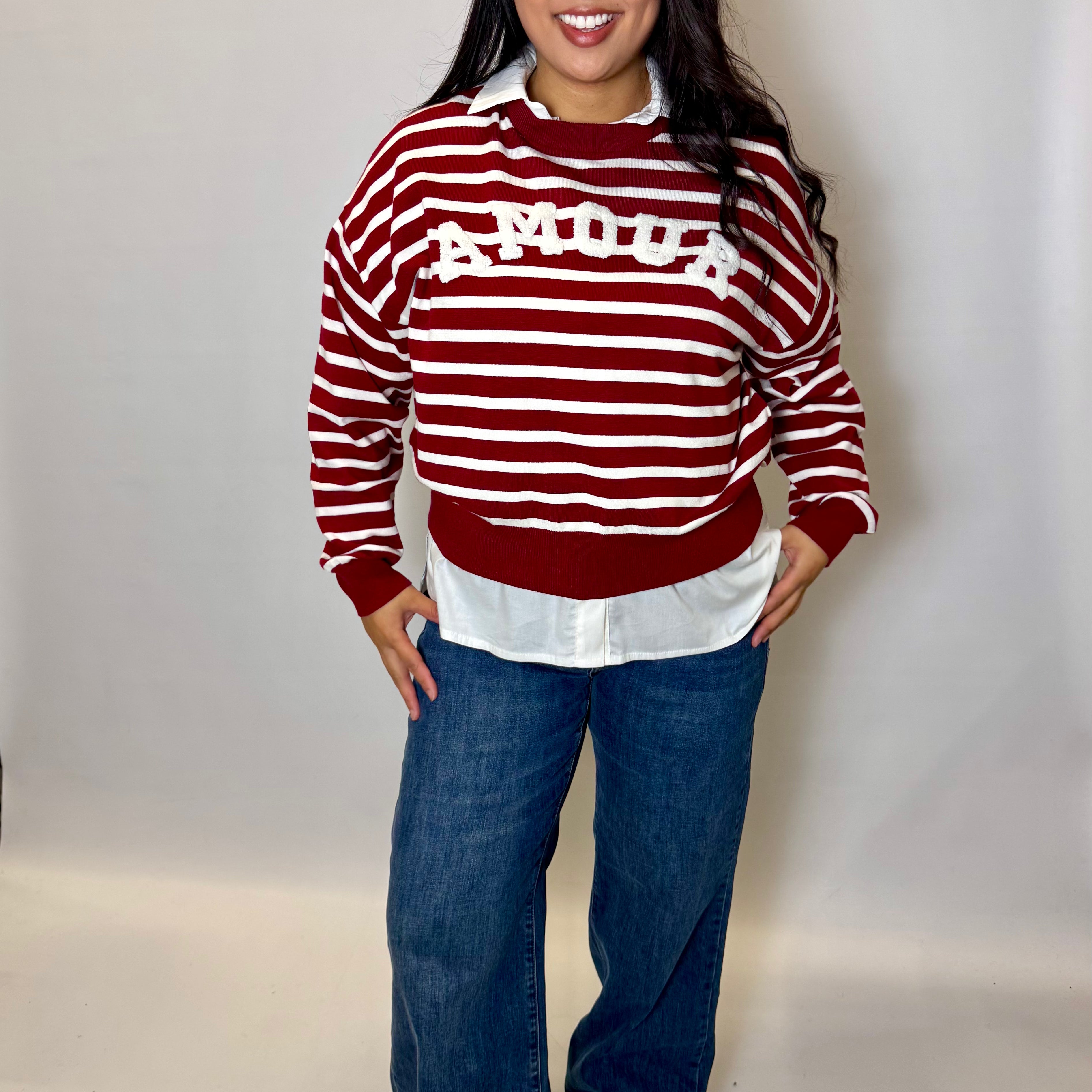 Woman wearing a red and white striped sweater with 'AMOUR' text, white shirt, blue jeans, and white shoes on a plain background.