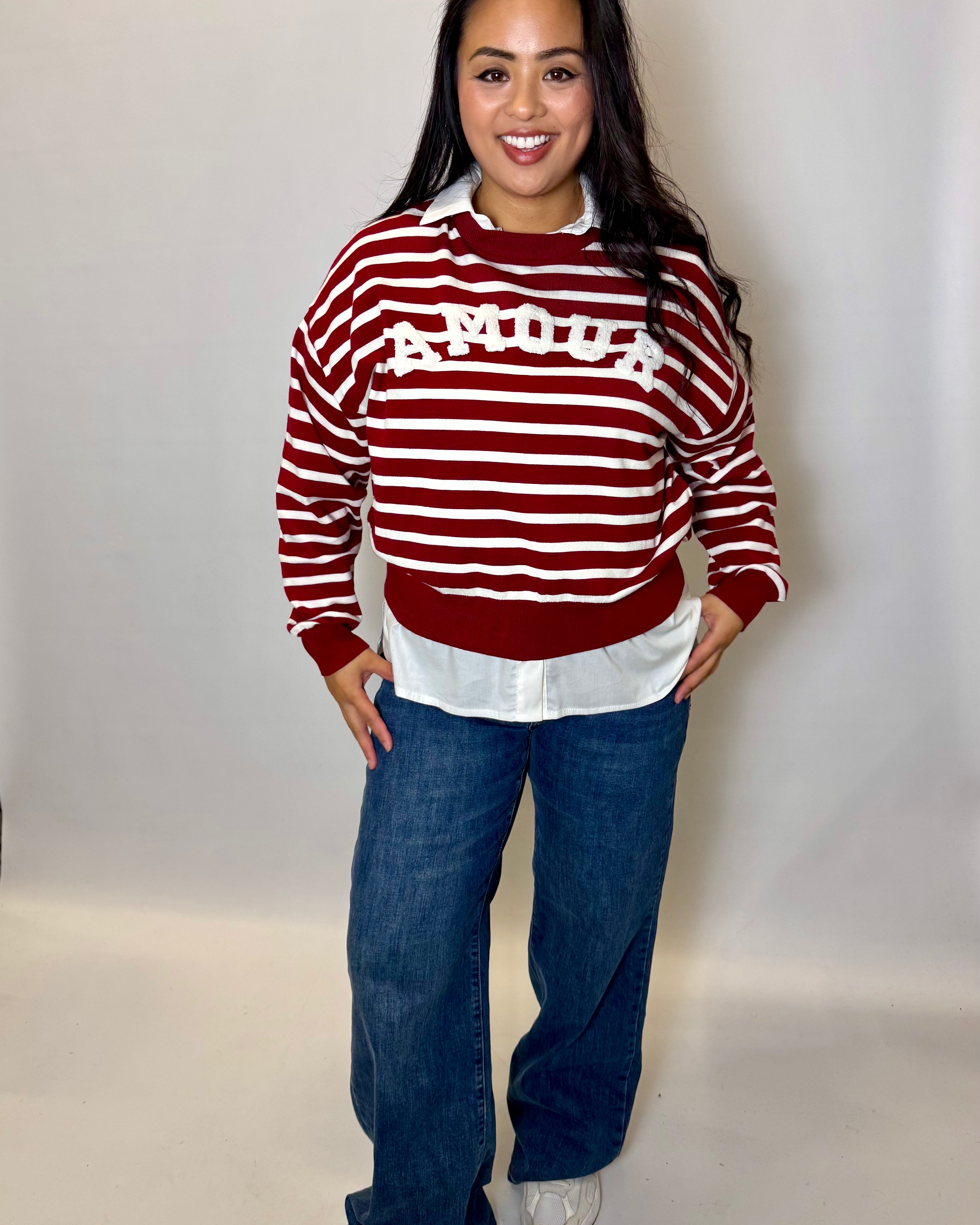 Woman wearing a red and white striped sweater with 'AMOUR' text, white shirt, blue jeans, and white shoes on a plain background.