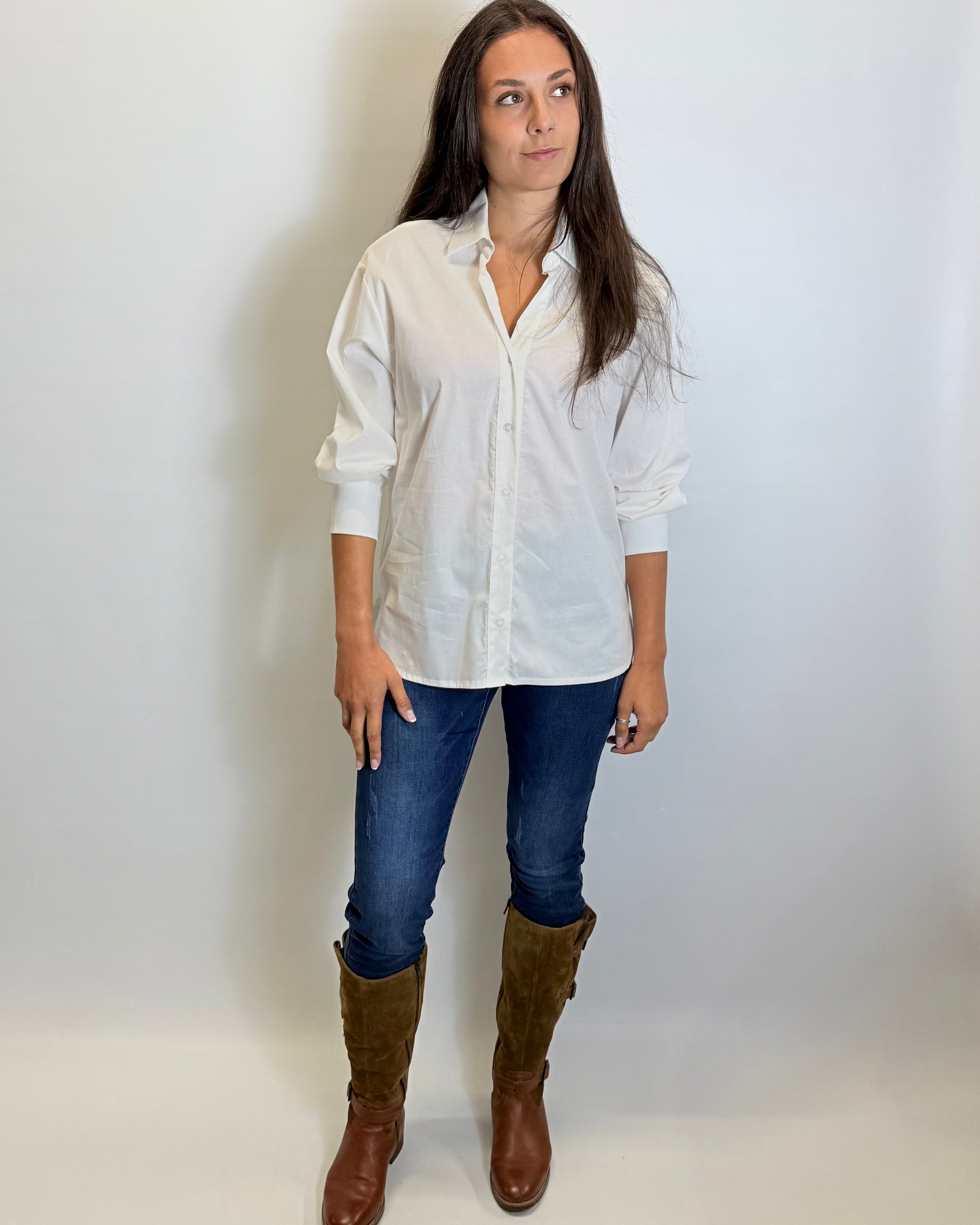 Woman wearing a white shirt, blue jeans, and brown boots against a plain background