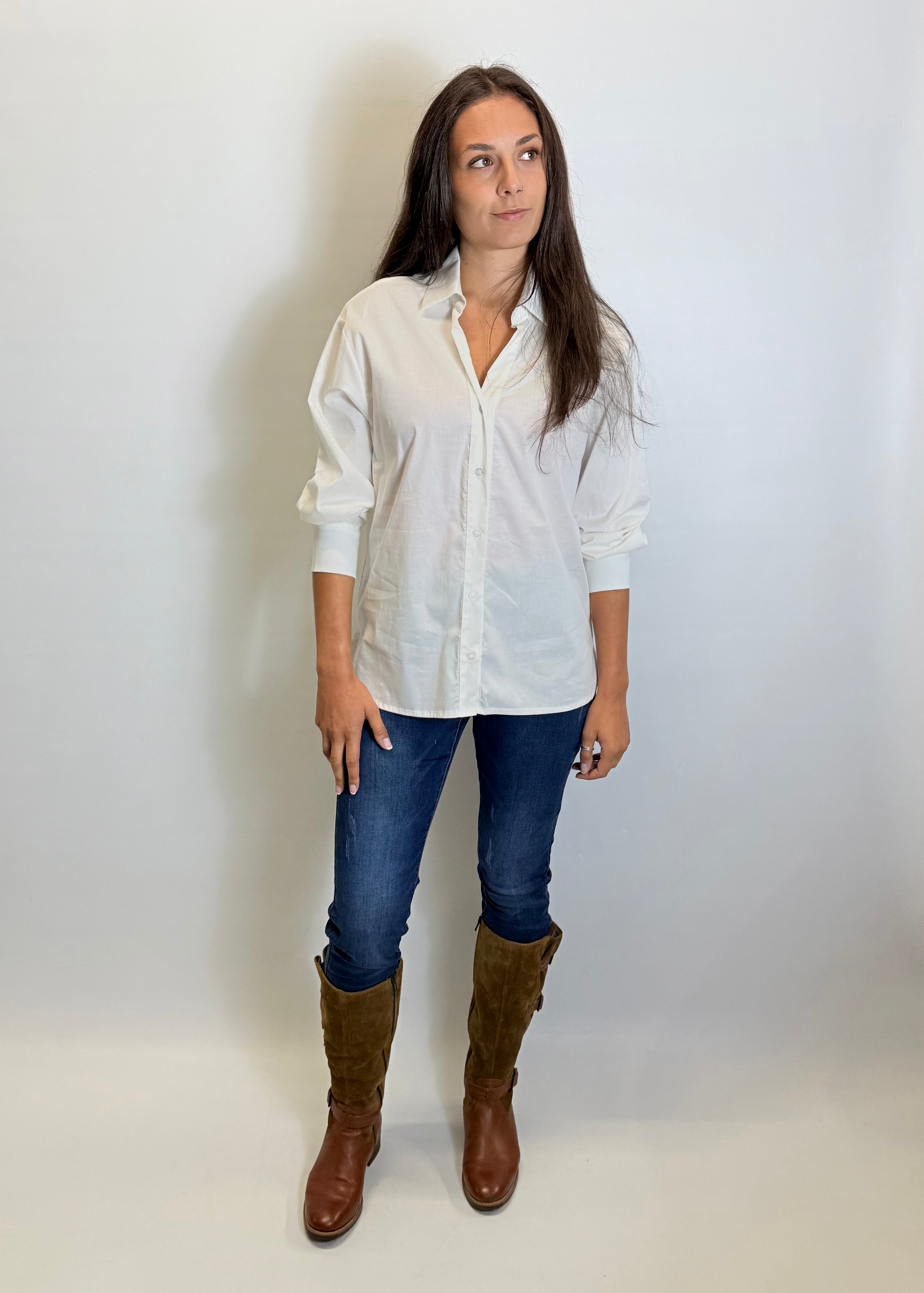 Woman wearing a white shirt, blue jeans, and brown boots against a plain background