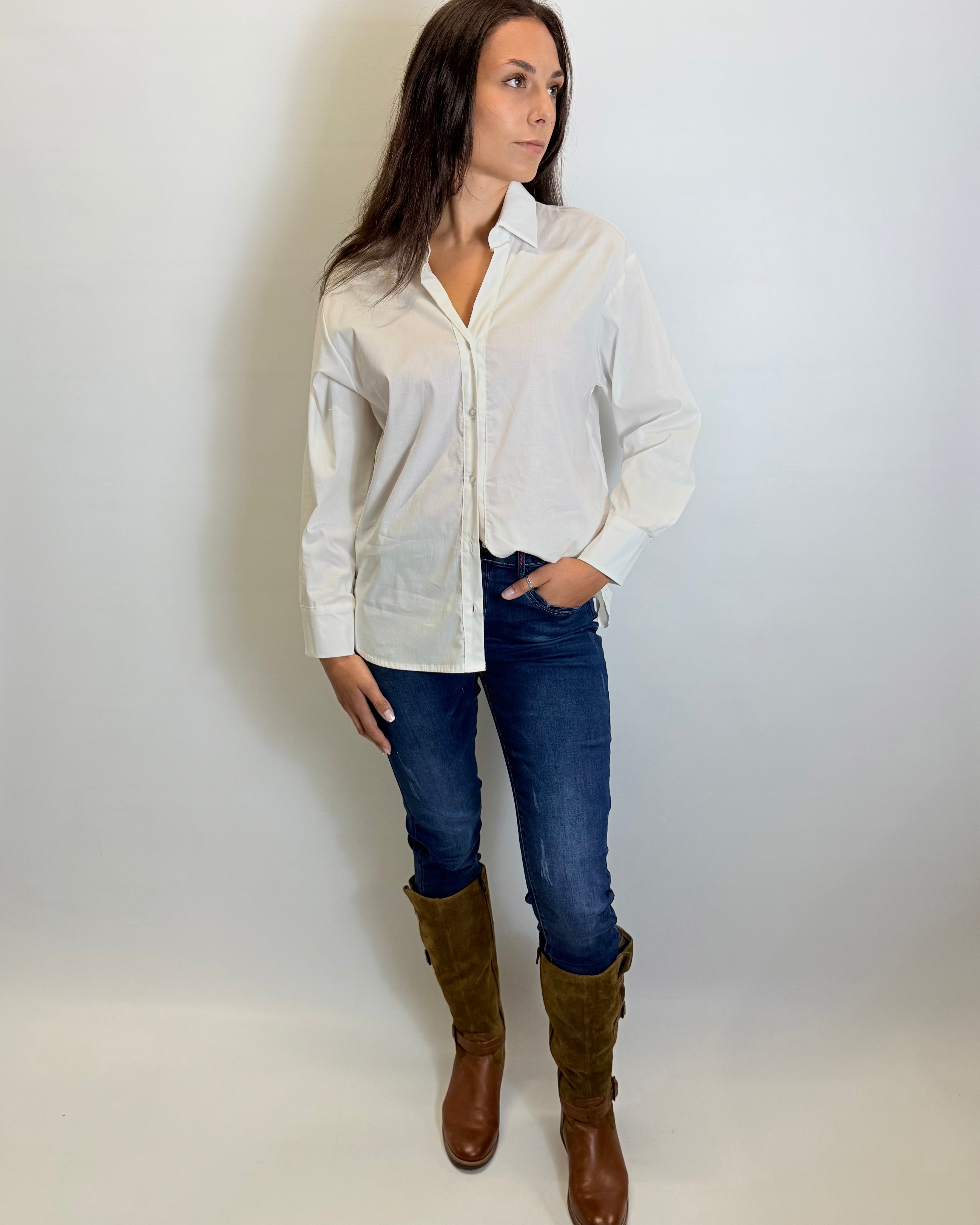 Woman wearing a white shirt, blue jeans, and brown boots against a plain background