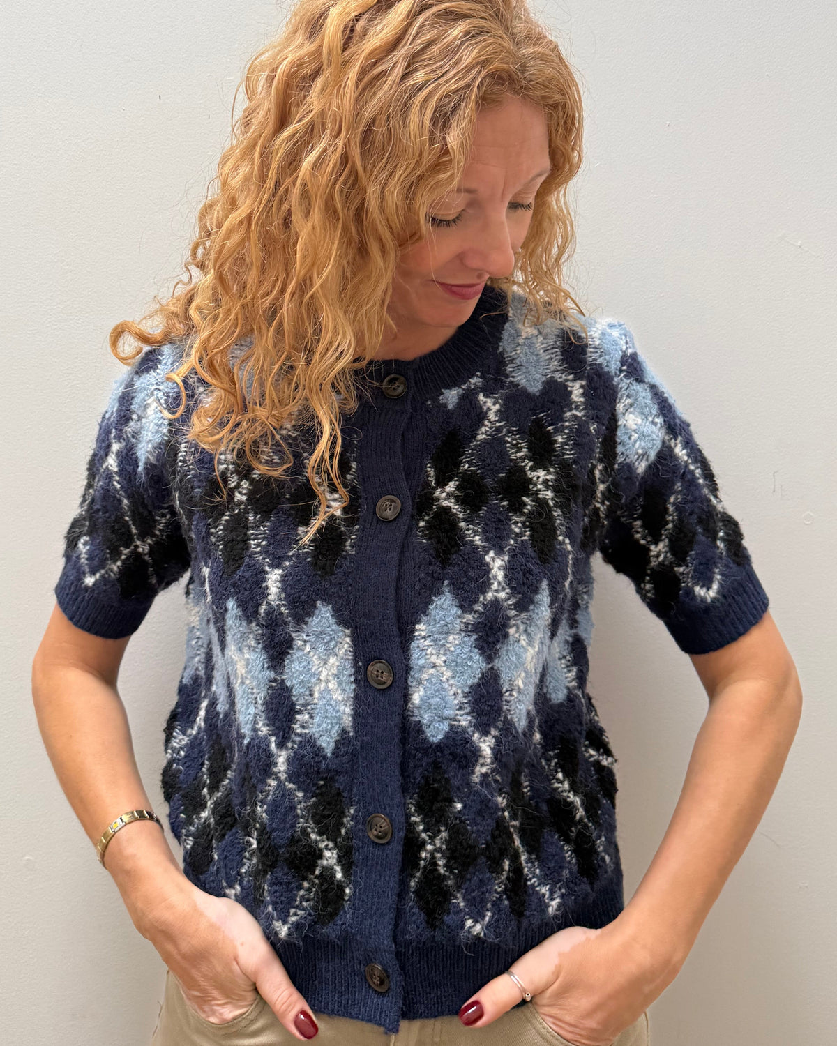 A woman with curly blonde hair wearing a navy blue short-sleeve button-up cardigan with a light blue and white argyle pattern, paired with beige pants, standing against a plain light gray wall.