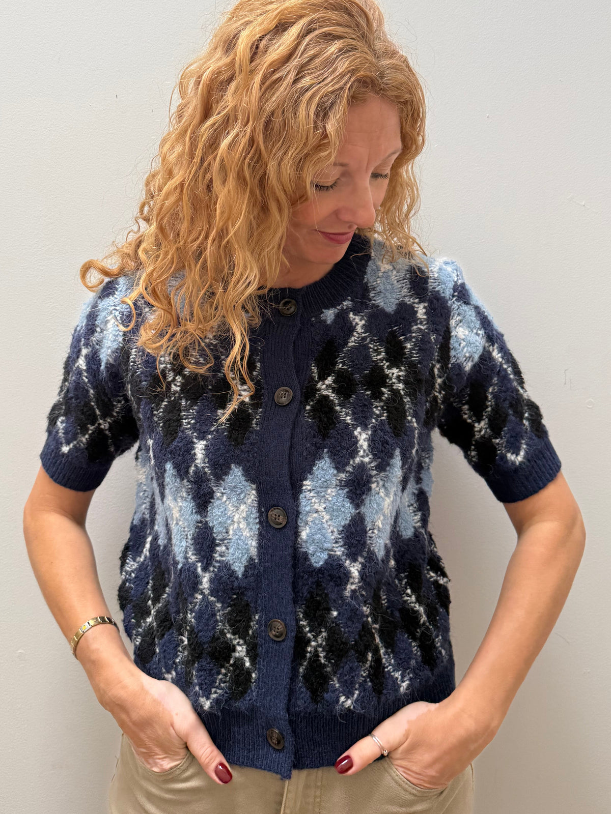 A woman with curly blonde hair wearing a navy blue short-sleeve button-up cardigan with a light blue and white argyle pattern, paired with beige pants, standing against a plain light gray wall.