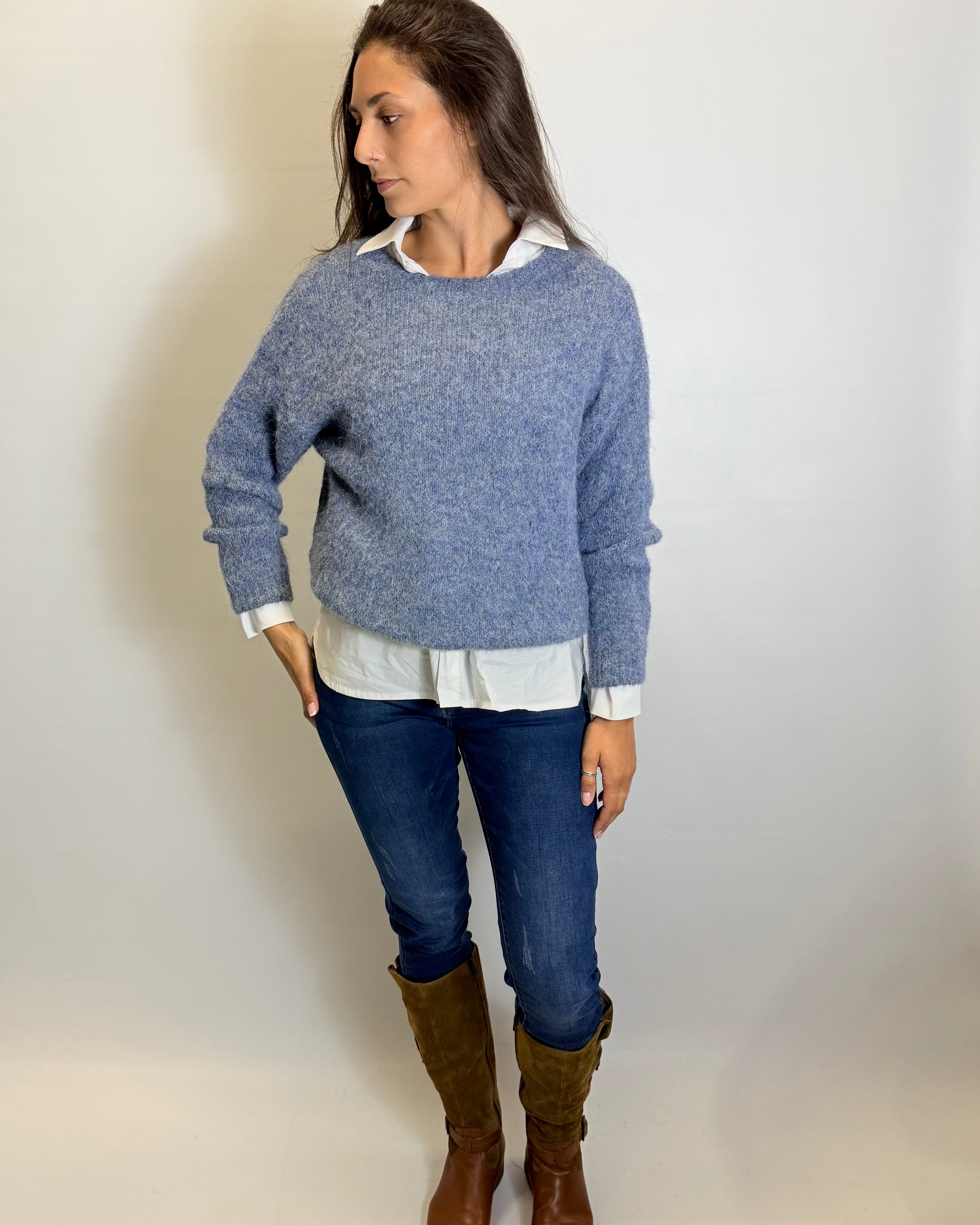 Woman wearing a blue sweater, white shirt, blue jeans, and brown boots against a plain background