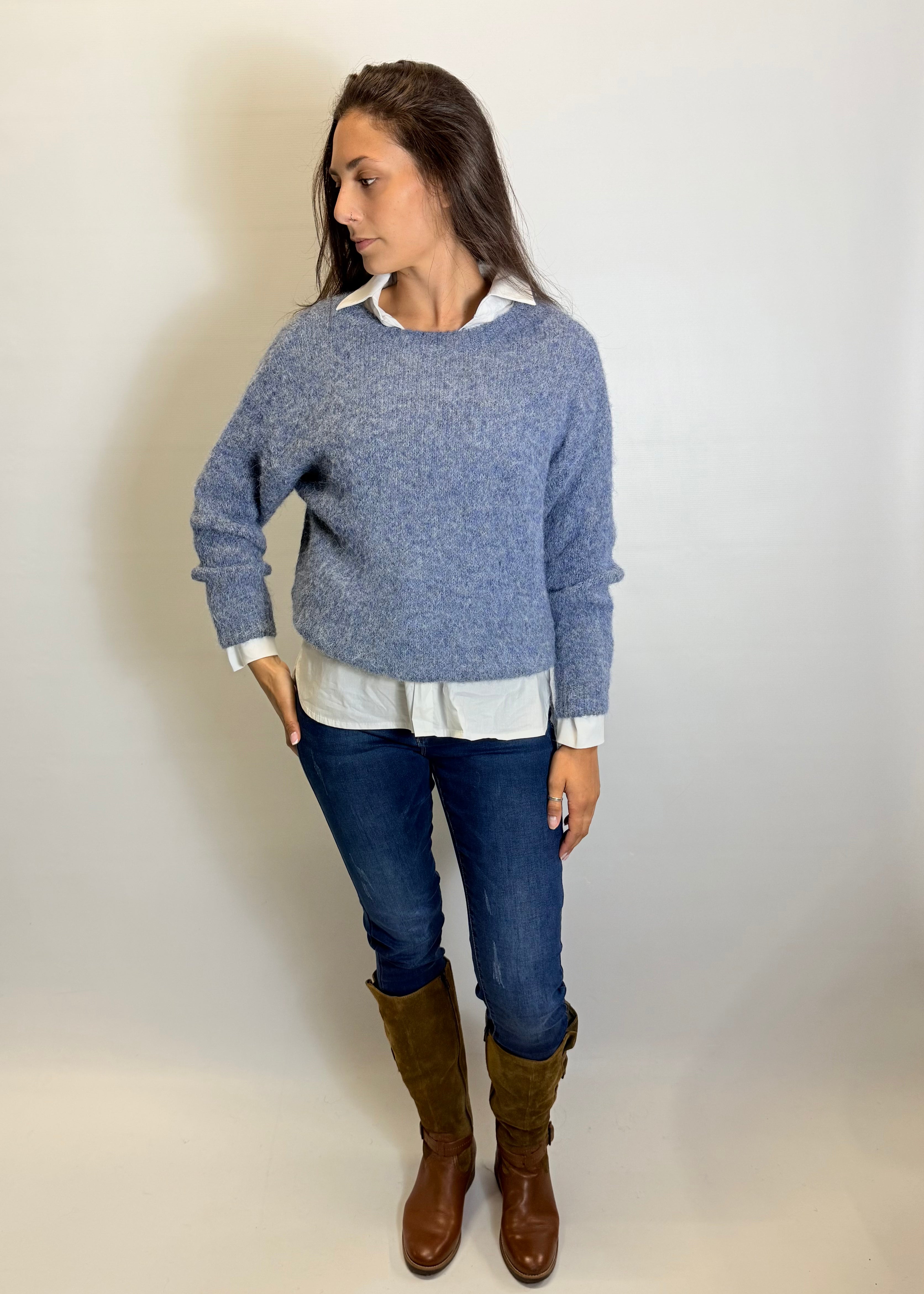 Woman wearing a blue sweater, white shirt, blue jeans, and brown boots against a plain background