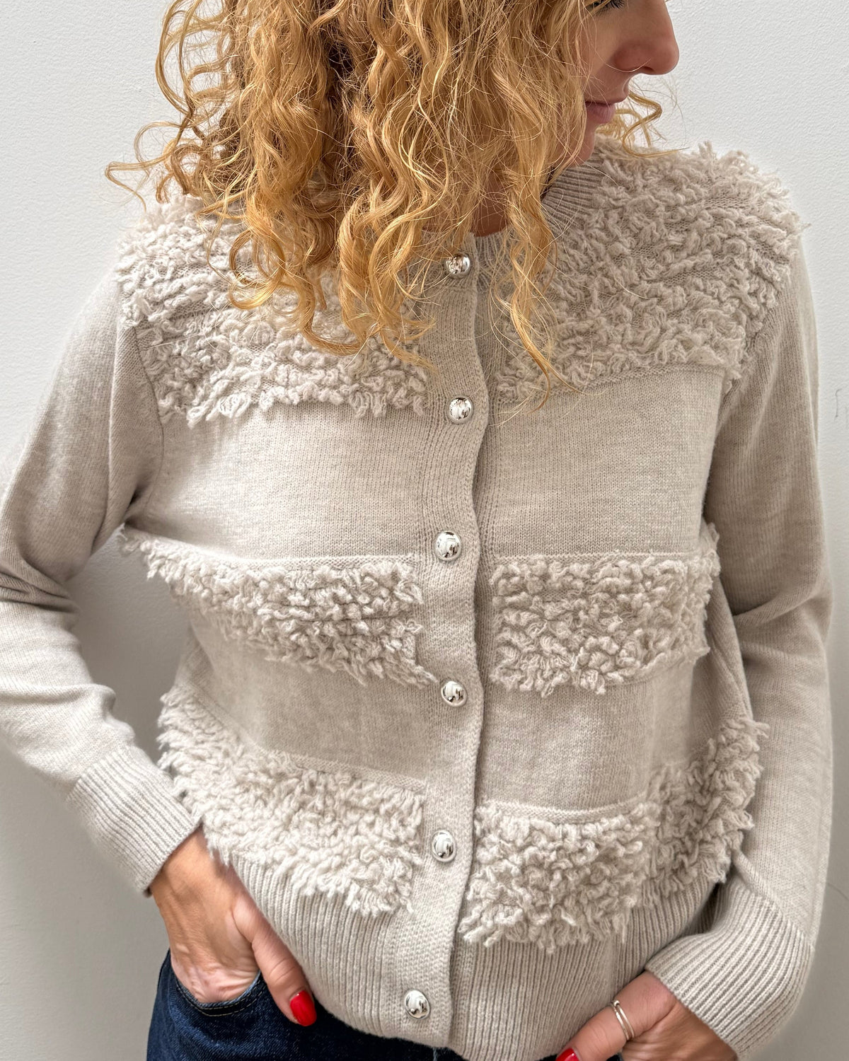 A woman with curly blonde hair wearing a beige textured knit cardigan with fuzzy trim and buttons, paired with dark blue jeans, standing against a plain light gray wall.