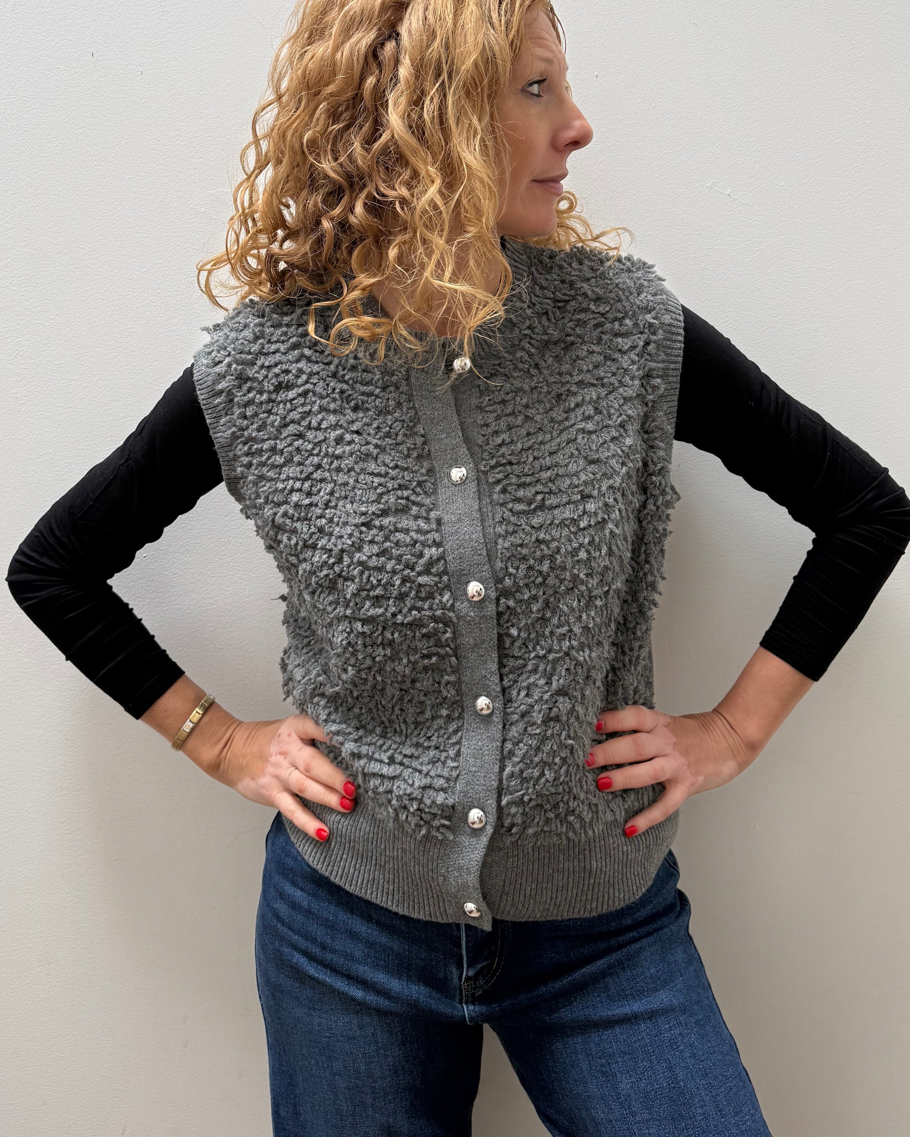 A woman with curly blonde hair models a gray textured sherpa button-up vest over a black long-sleeve shirt and blue jeans, standing against a plain light gray wall.