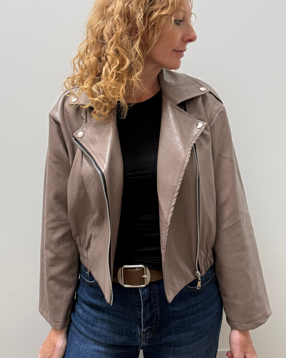 A woman with curly blonde hair wearing a taupe faux-leather moto jacket, black top, and blue jeans, standing against a plain light gray wall.
