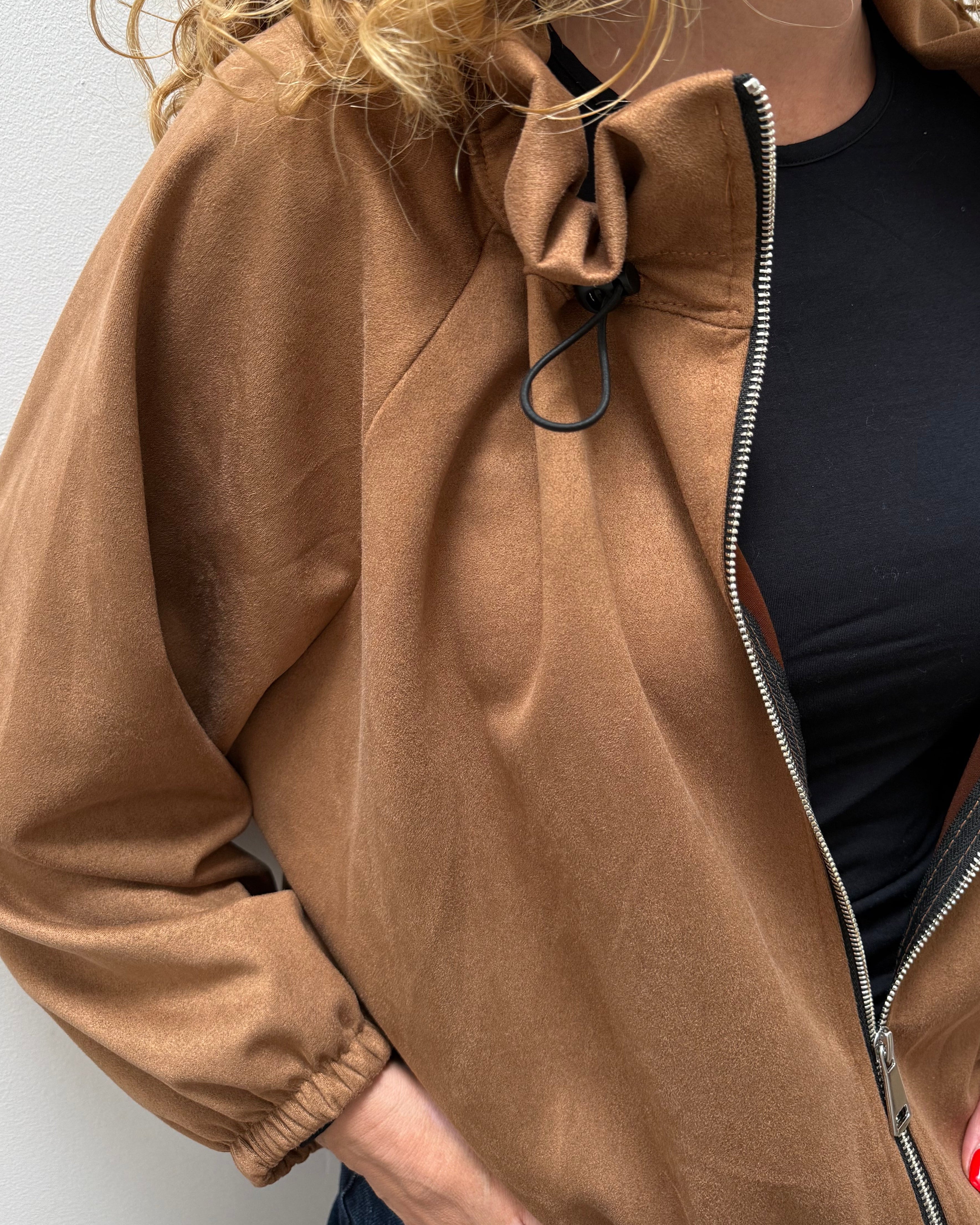 A close-up of a woman wearing a camel brown zip-up jacket over a black top, with her hands in her pockets against a plain light gray wall.