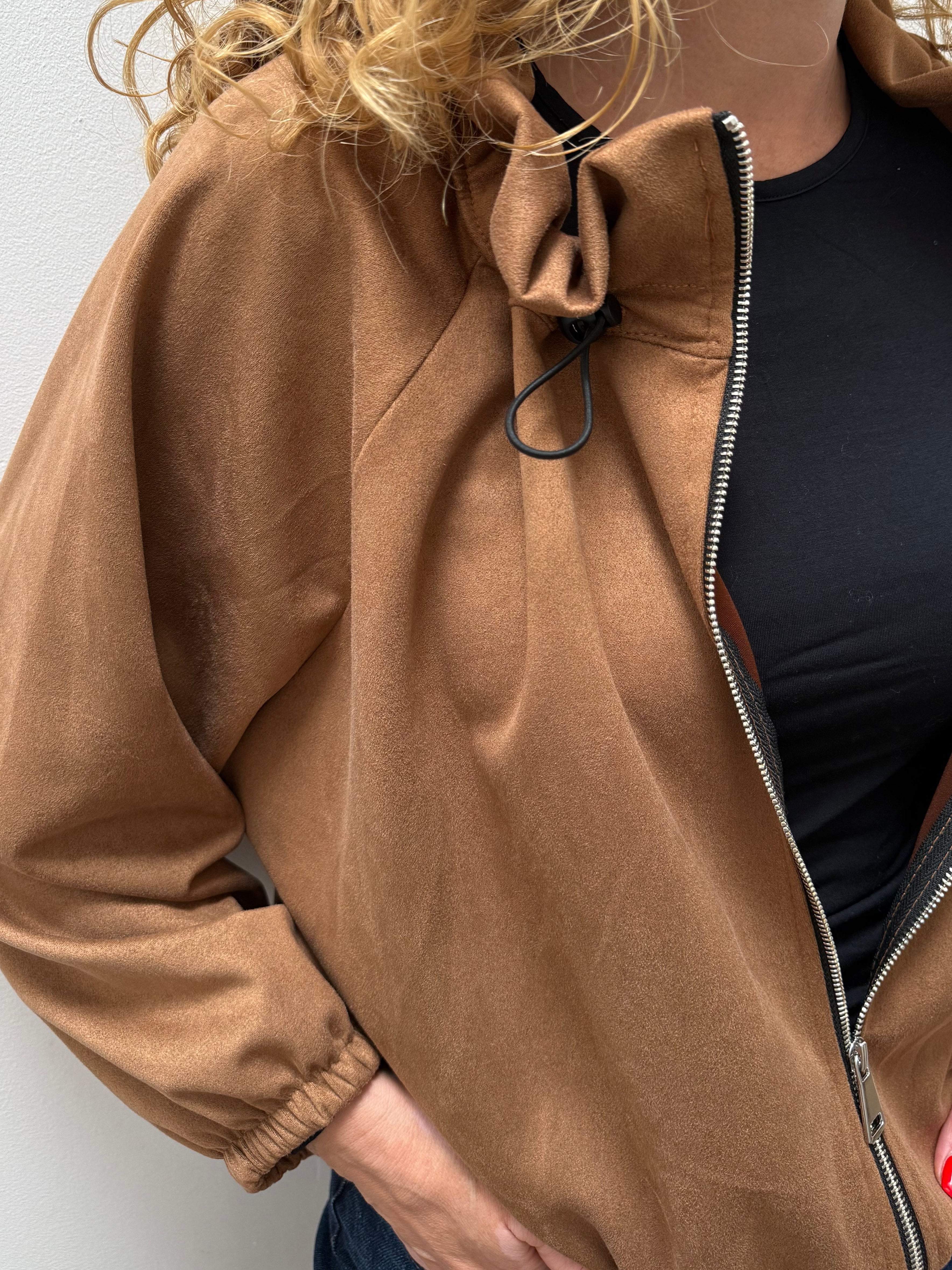 A close-up of a woman wearing a camel brown zip-up jacket over a black top, with her hands in her pockets against a plain light gray wall.