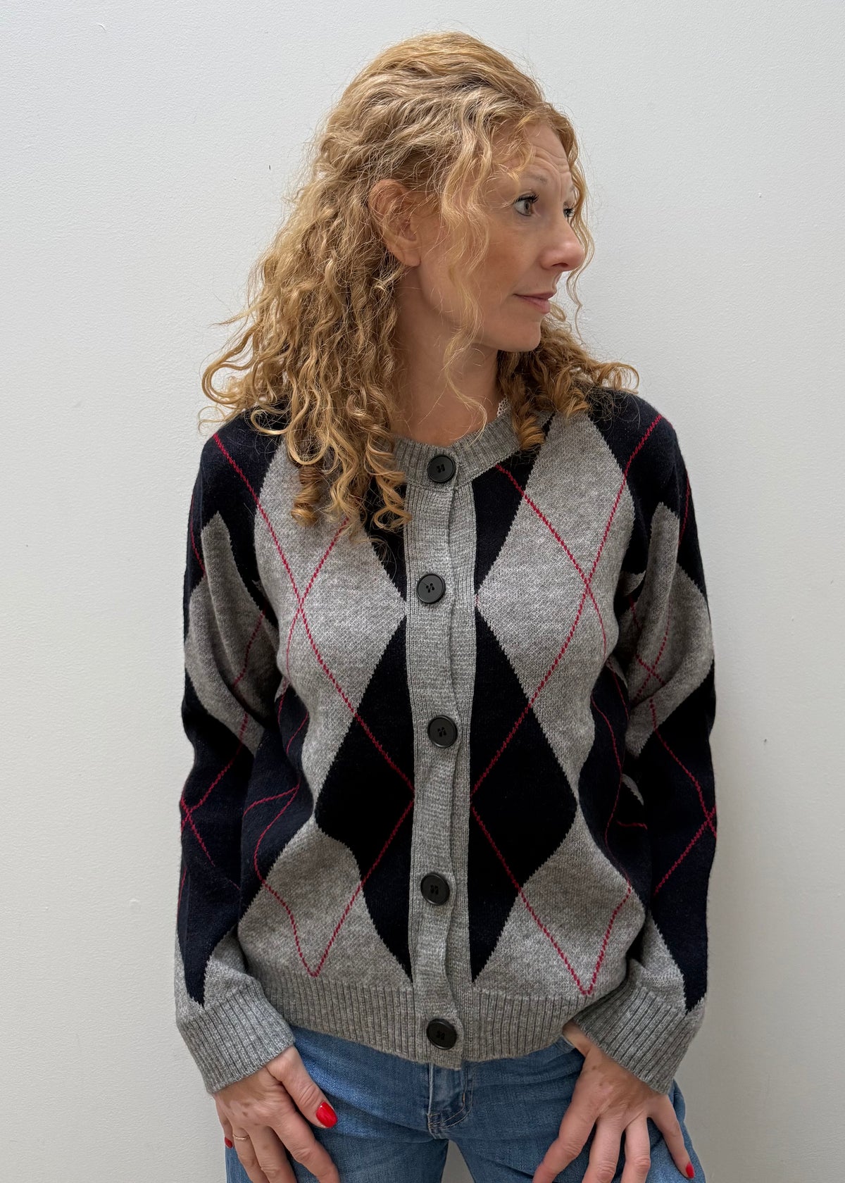 A woman with curly blonde hair wearing a gray, navy, and red argyle cardigan sweater with black buttons, paired with blue jeans, standing against a plain light gray wall.