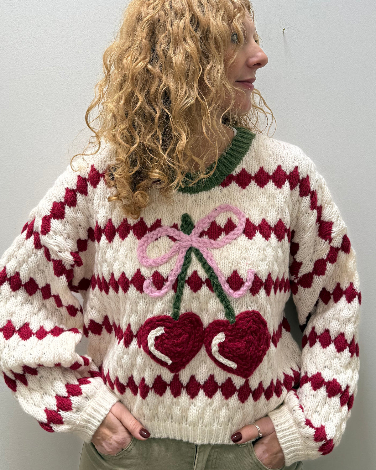 A woman with curly blonde hair wearing a chunky knit sweater with a red and white diamond pattern and a large crocheted cherry design on the front, paired with khaki pants, standing against a plain light gray wall.