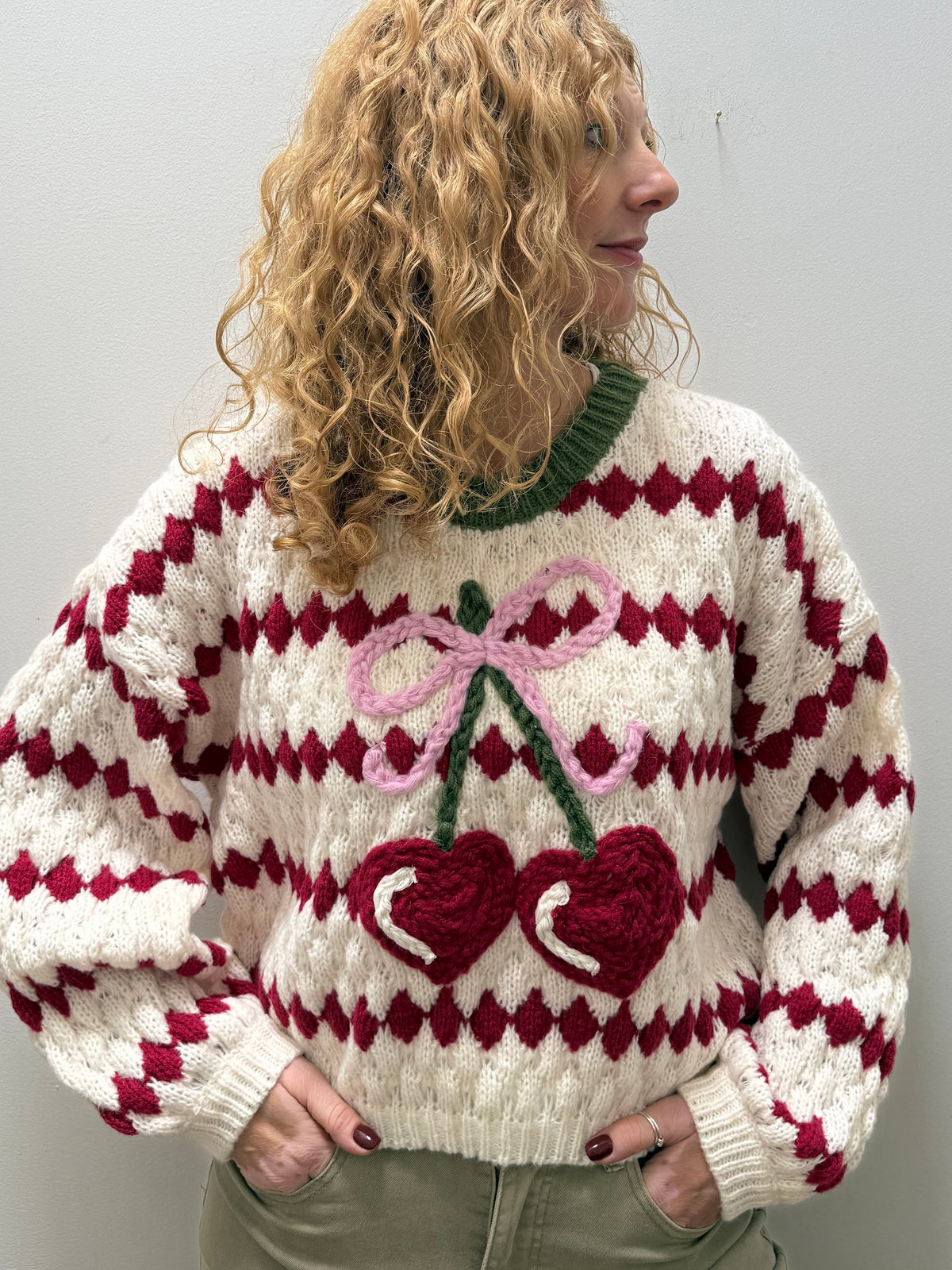 A woman with curly blonde hair wearing a chunky knit sweater with a red and white diamond pattern and a large crocheted cherry design on the front, paired with khaki pants, standing against a plain light gray wall.