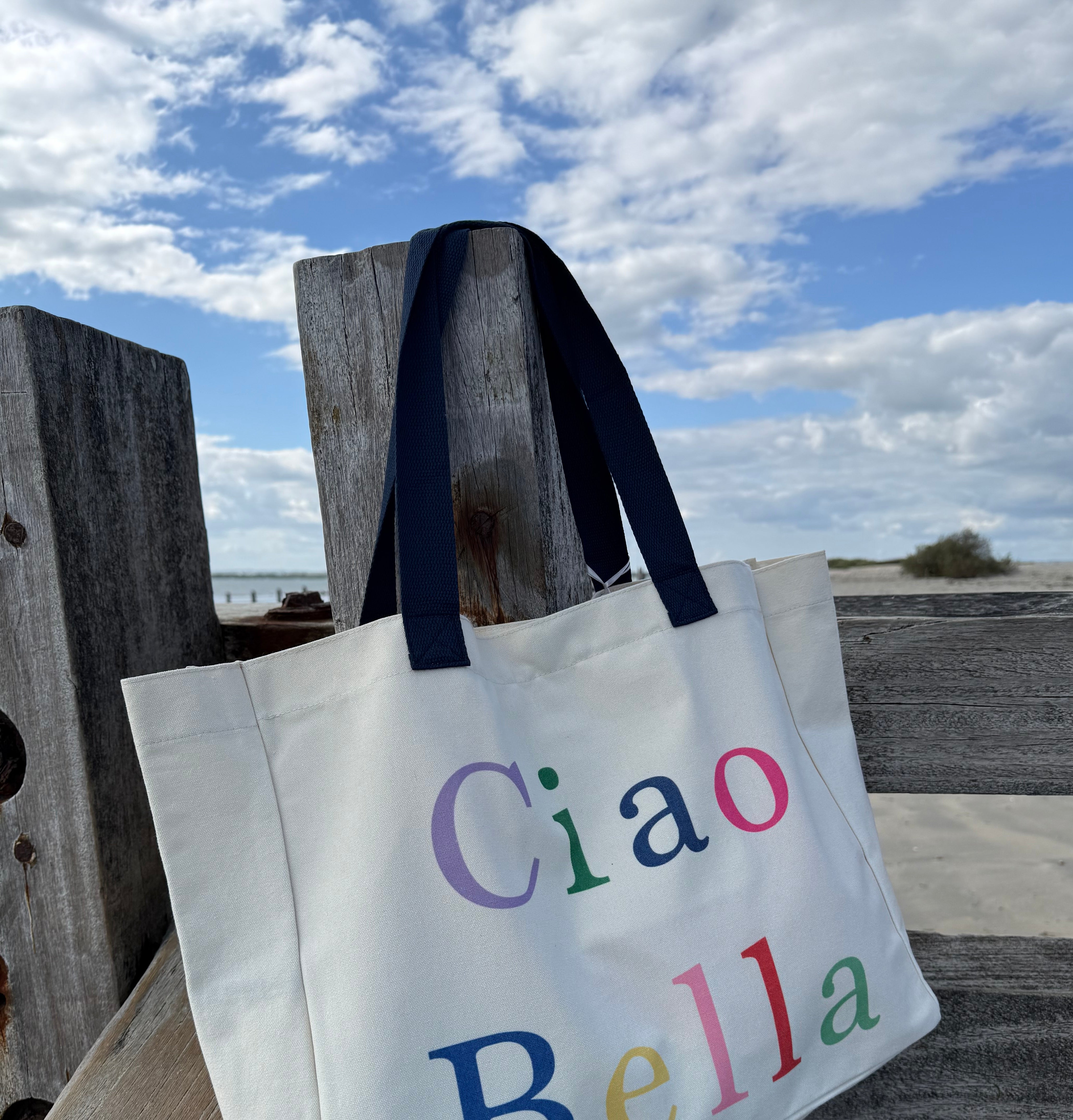 Tote bag with 'Ciao Bella' text on a wooden post against a cloudy sky.