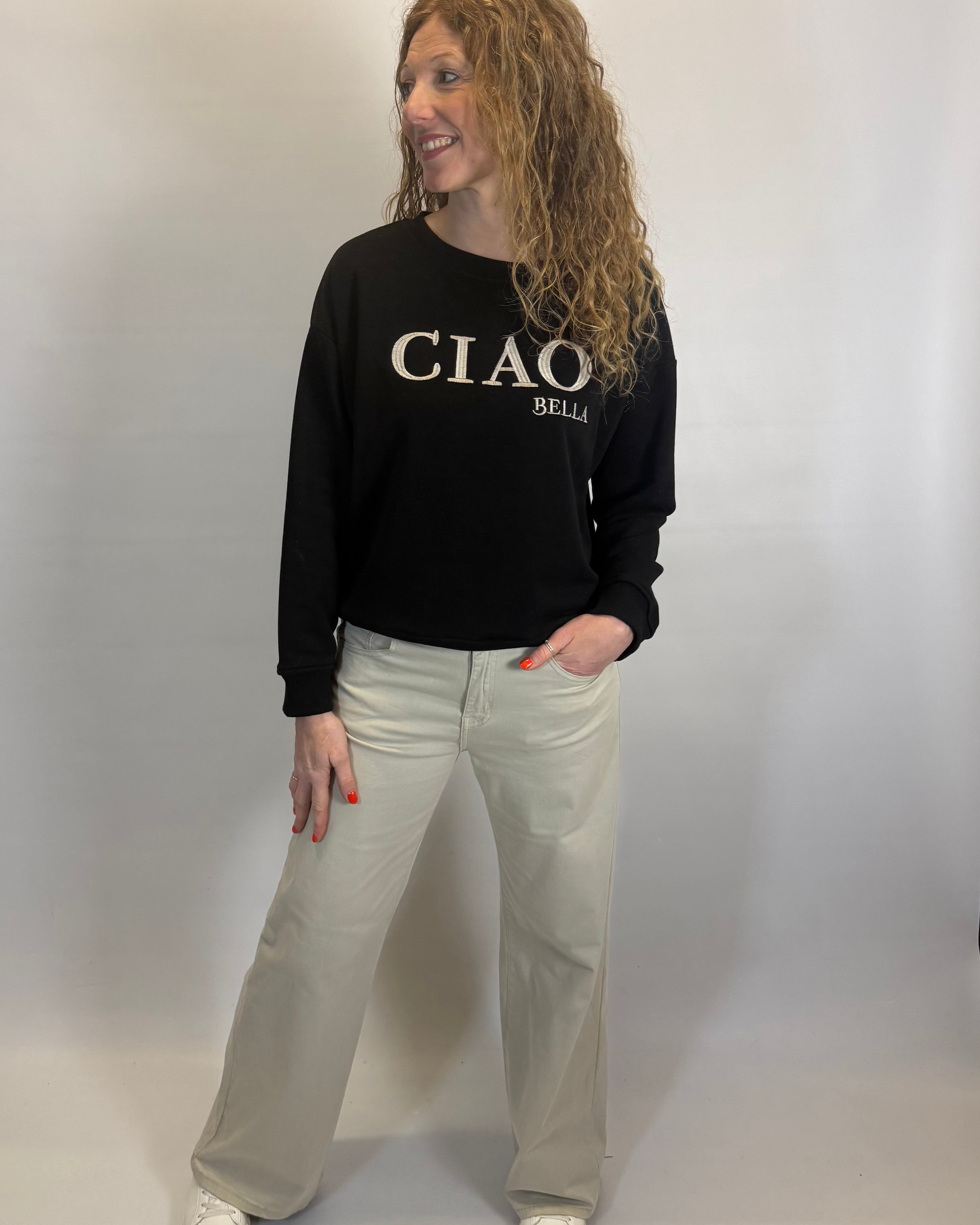 Person wearing a black sweatshirt with 'CIAO BELLI' text and light-colored pants on a plain background