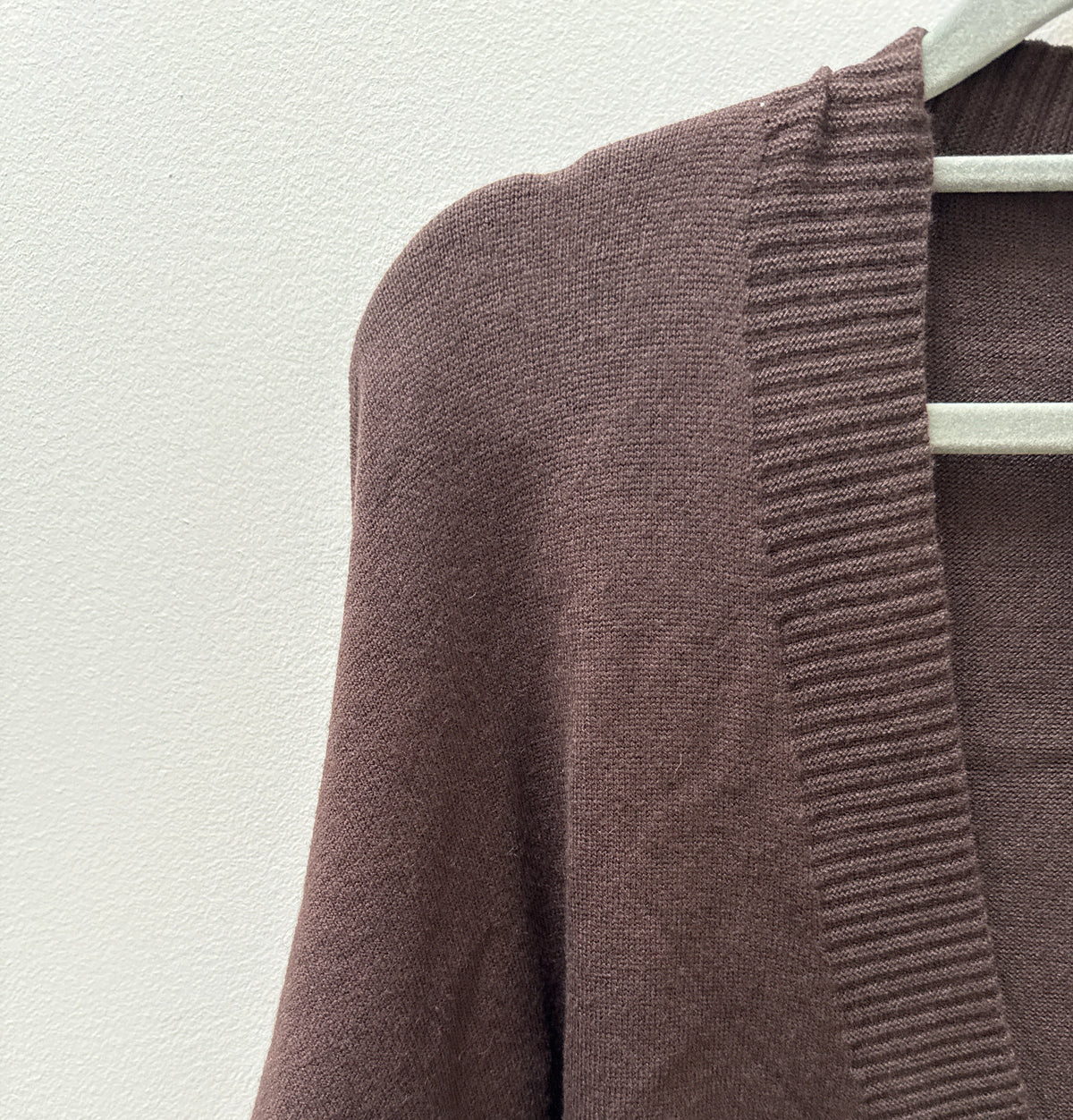 A close-up of a brown knit cardigan sweater hanging on a white hanger against a textured off-white wall. The cardigan features a ribbed V-neck trim and a single visible light-colored button.