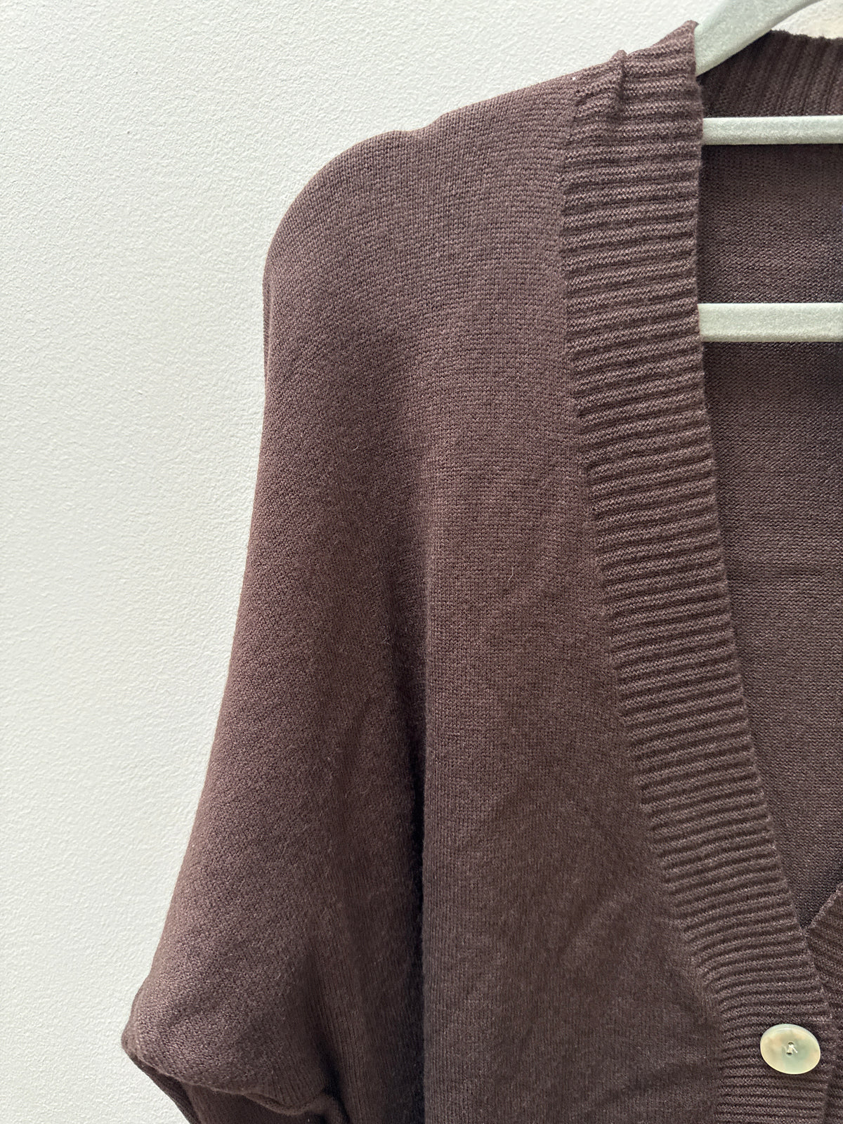 A close-up of a brown knit cardigan sweater hanging on a white hanger against a textured off-white wall. The cardigan features a ribbed V-neck trim and a single visible light-colored button.