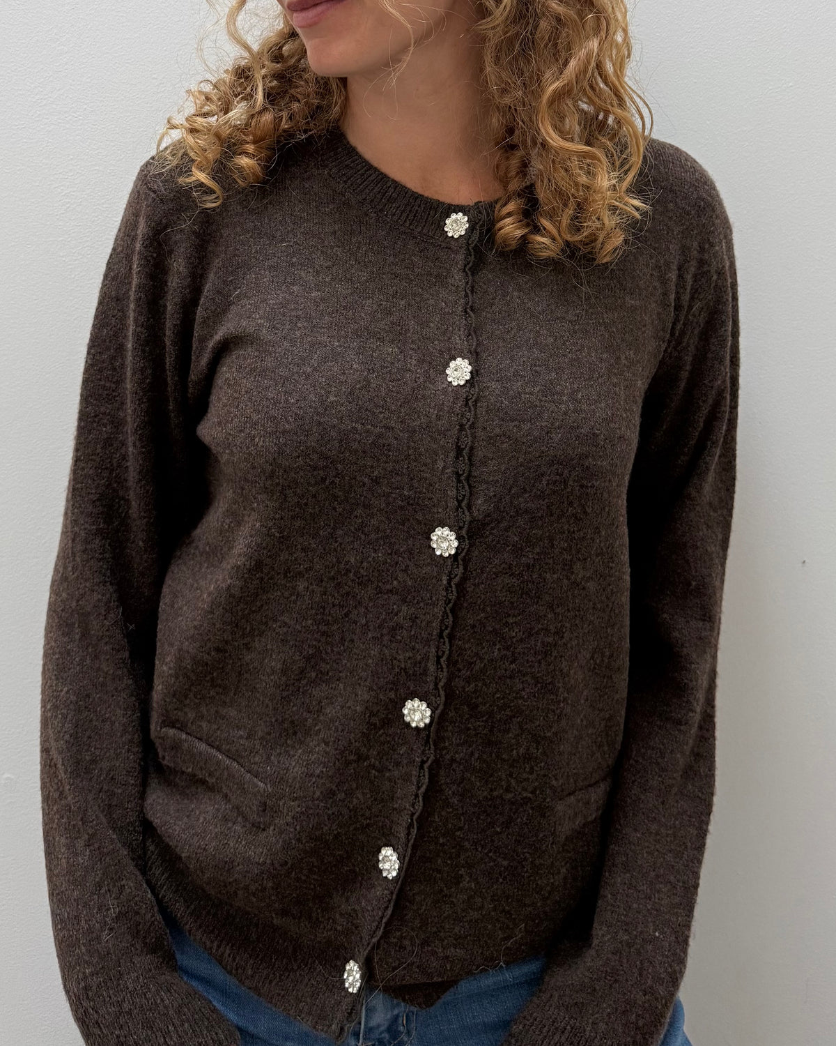 A woman wearing a dark brown knit cardigan sweater with decorative white buttons, paired with blue jeans, shown against a plain light gray wall.