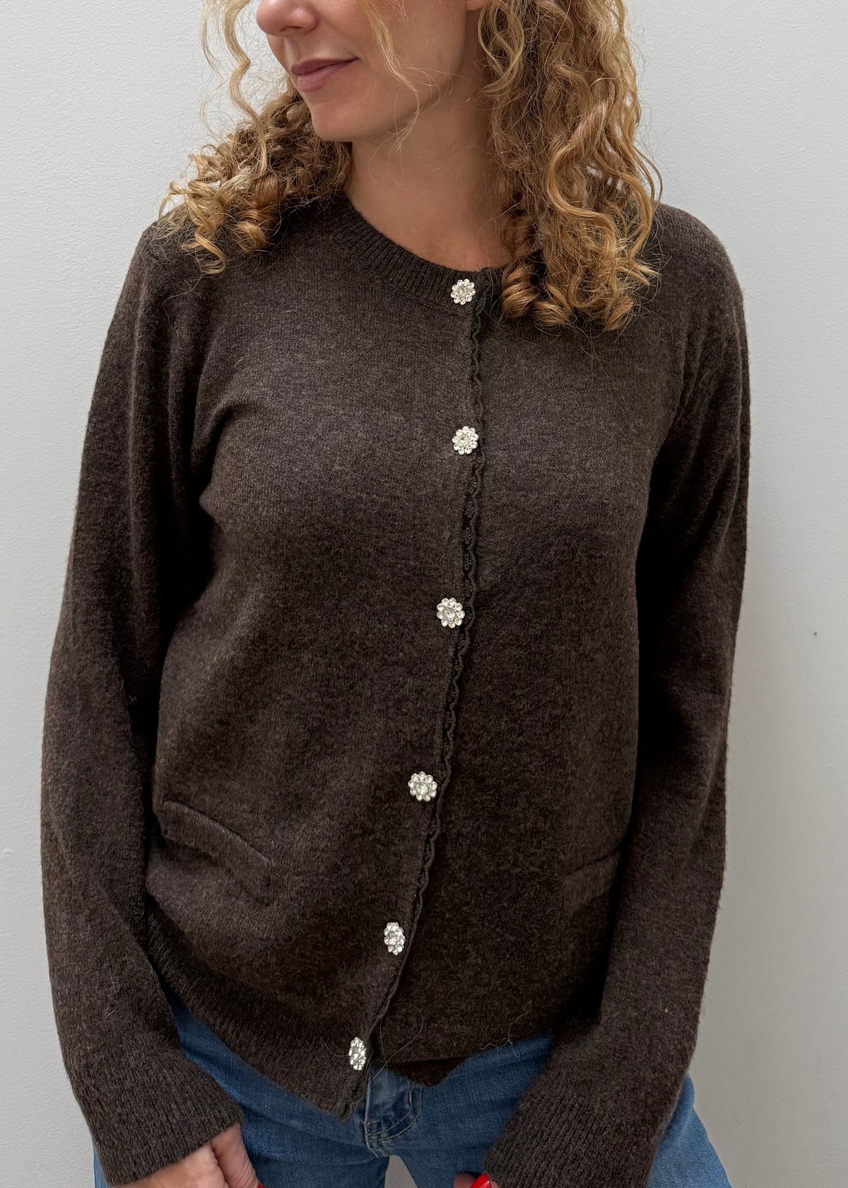 A woman wearing a dark brown knit cardigan sweater with decorative white buttons, paired with blue jeans, shown against a plain light gray wall.