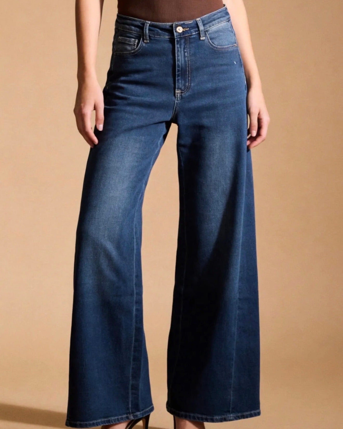 A model wearing dark blue wide-leg denim jeans with a brown bodysuit and black high-heeled sandals, standing against a solid beige studio background.