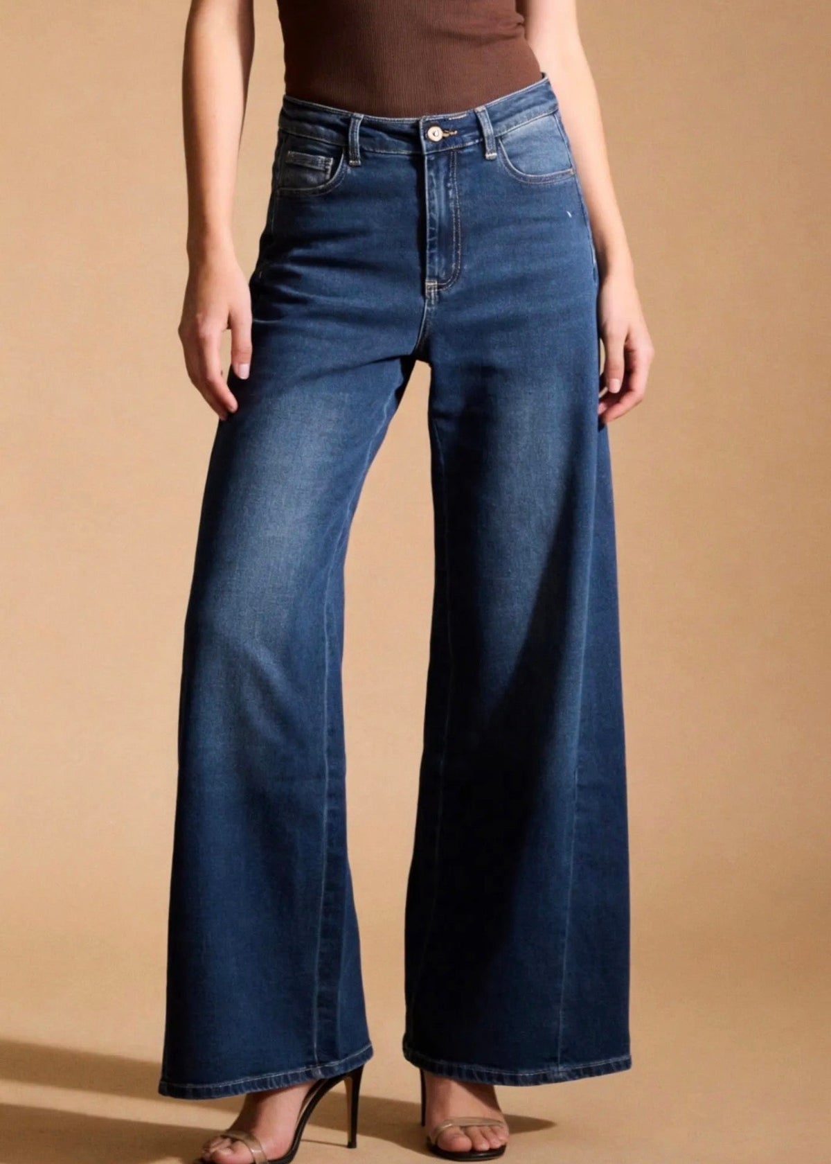 A model wearing dark blue wide-leg denim jeans with a brown bodysuit and black high-heeled sandals, standing against a solid beige studio background.