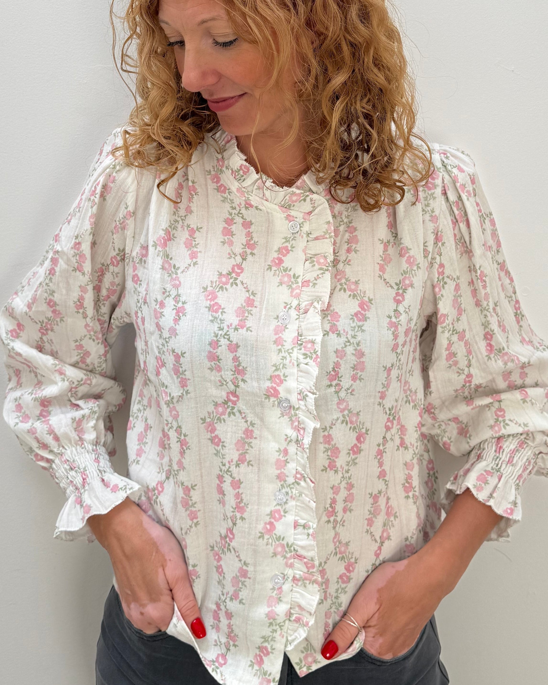 A woman with curly light brown hair wearing a cream-colored long-sleeve blouse with a pink floral pattern and dark gray pants, standing against a plain light gray wall.