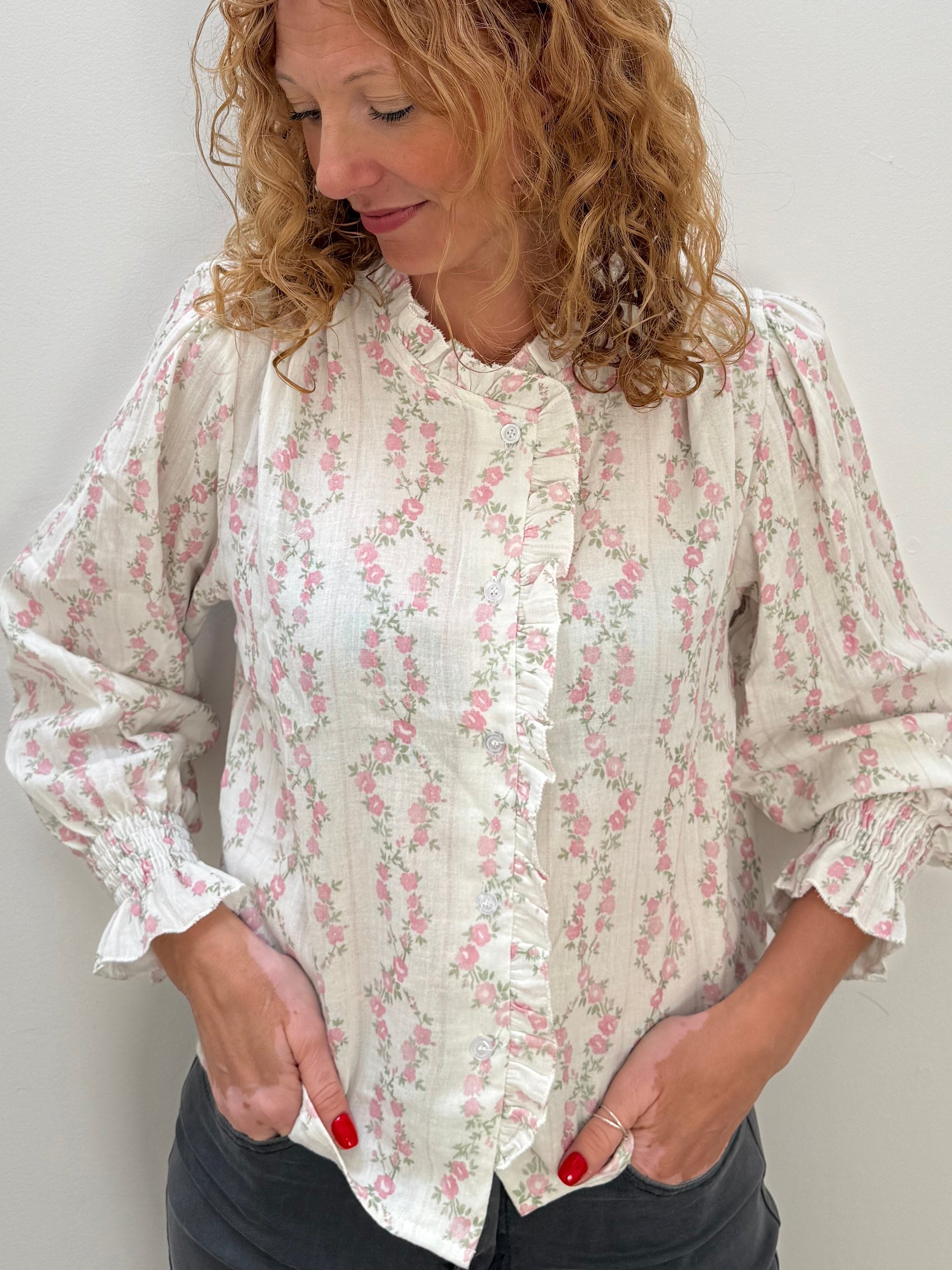 A woman with curly light brown hair wearing a cream-colored long-sleeve blouse with a pink floral pattern and dark gray pants, standing against a plain light gray wall.