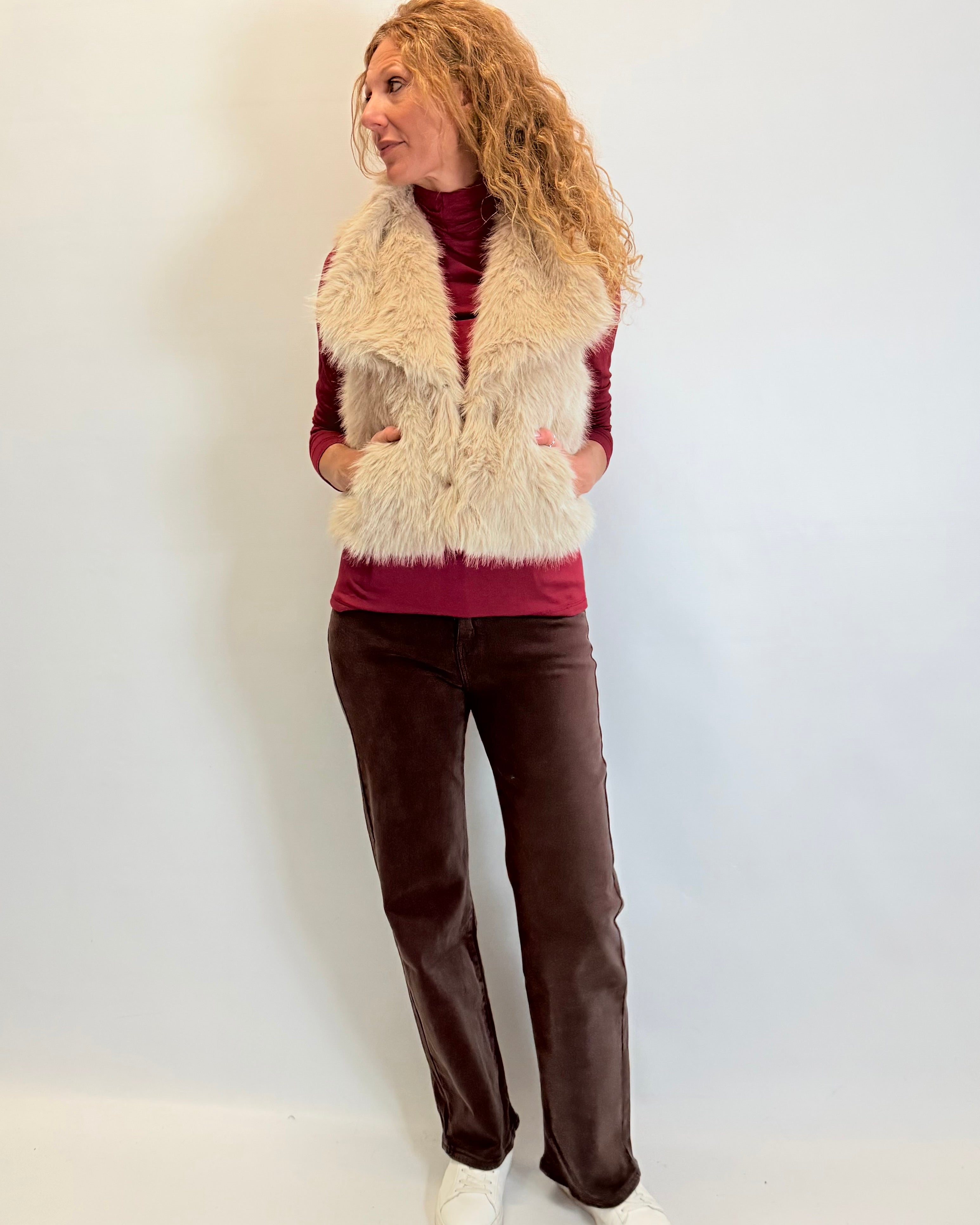 Woman wearing a fur vest, red top, brown pants, and white shoes against a plain background