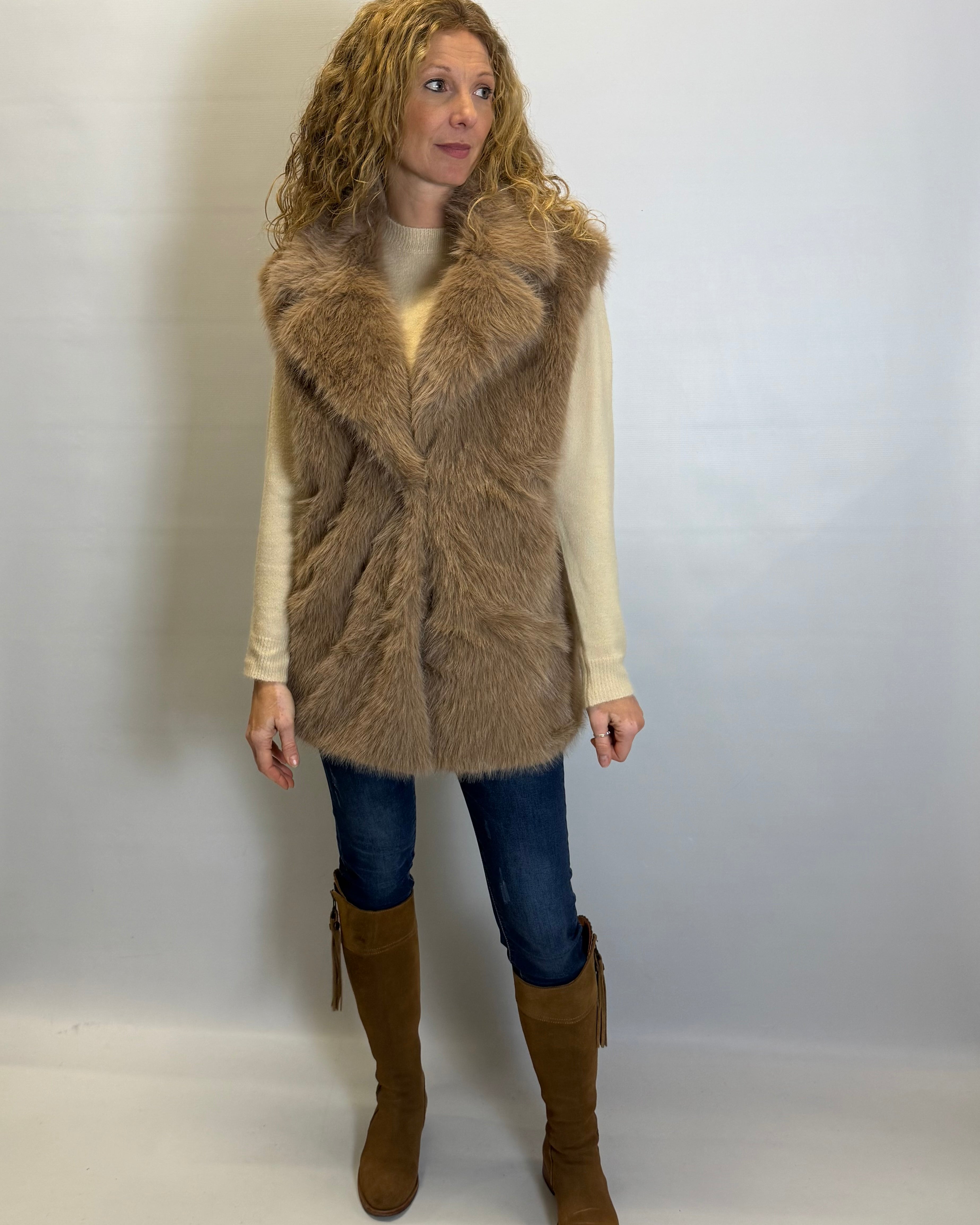 Woman wearing a brown fur vest, white shirt, blue jeans, and brown boots against a plain background