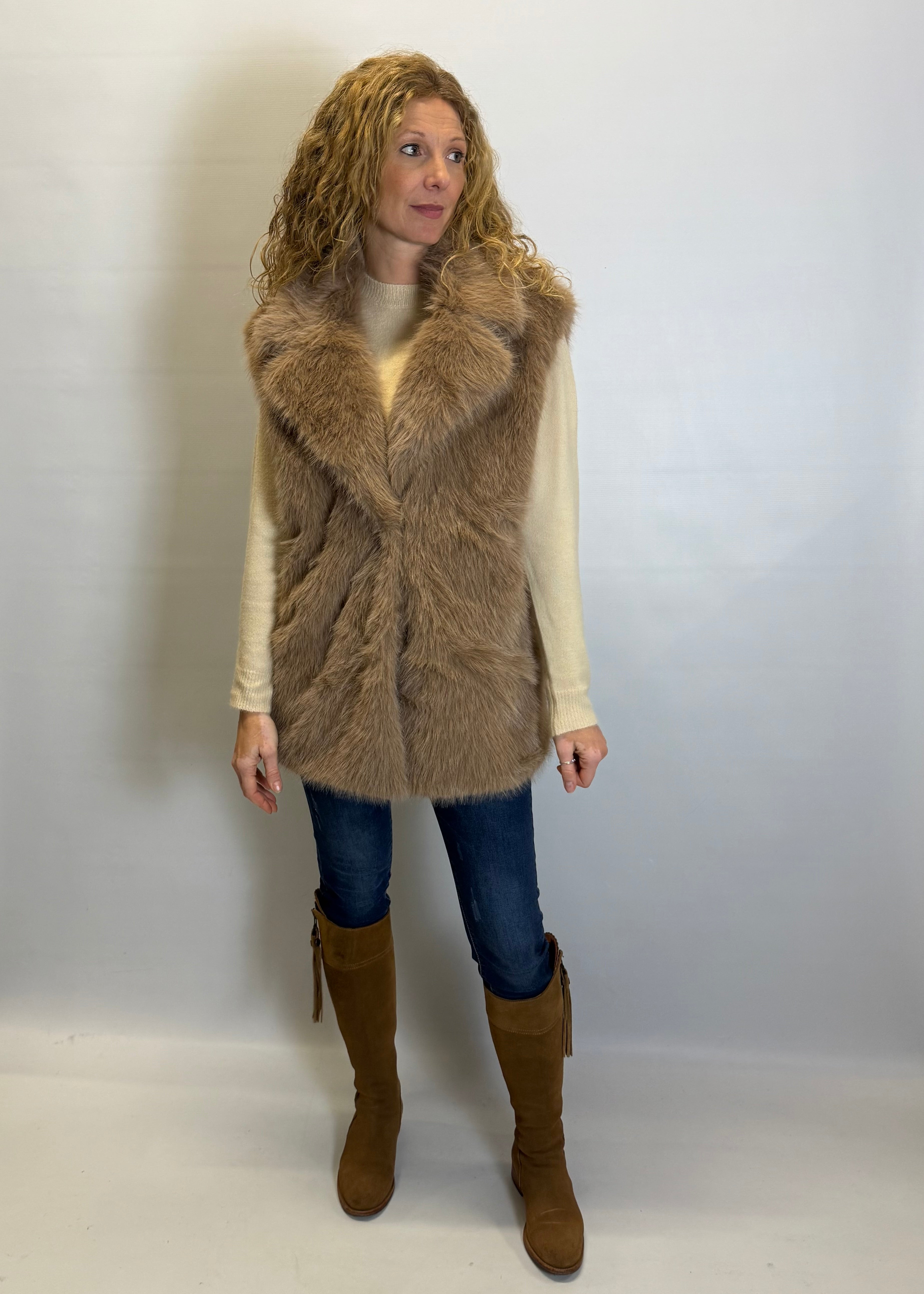 Woman wearing a brown fur vest, white shirt, blue jeans, and brown boots against a plain background