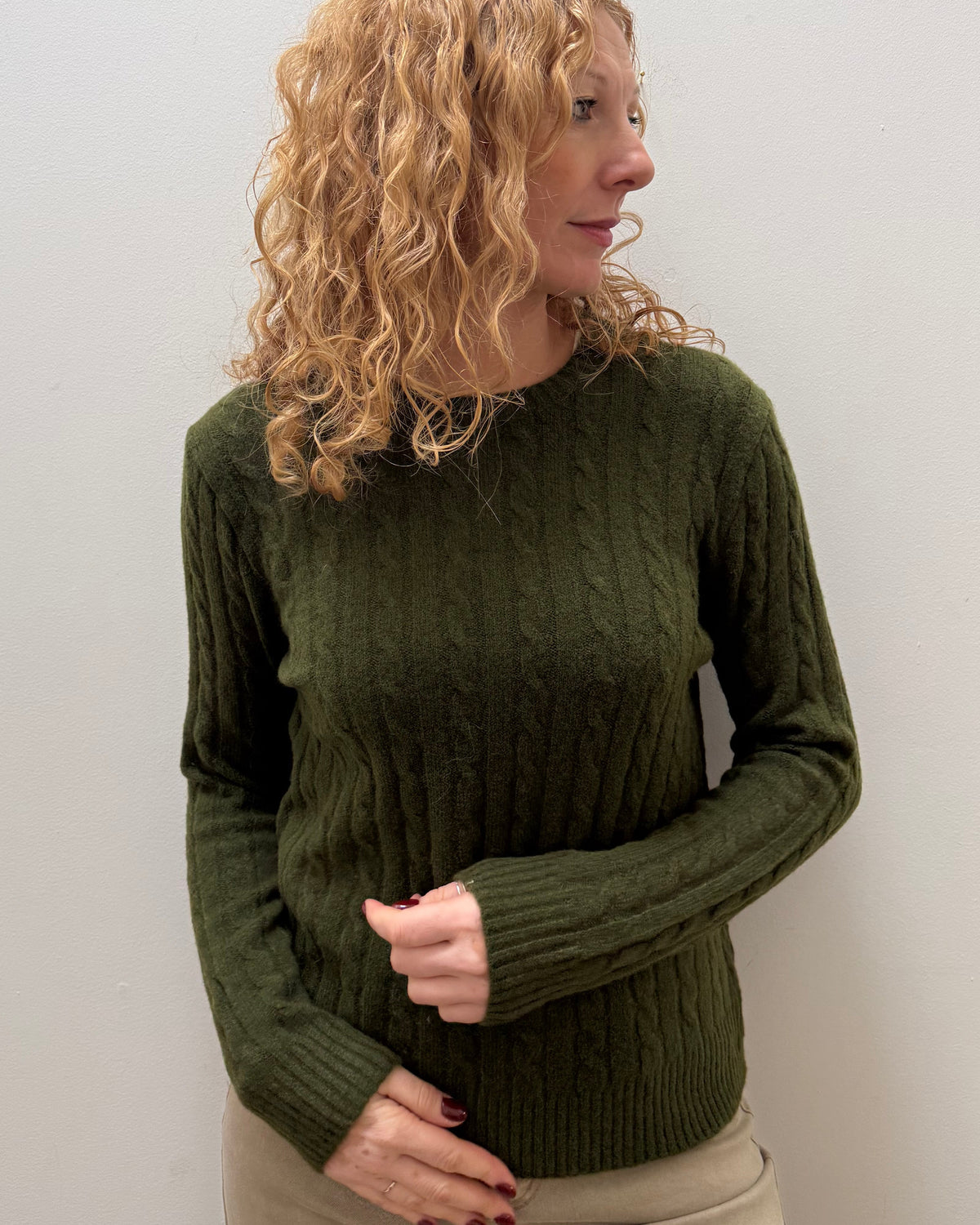 A woman with curly blonde hair wearing an olive green cable-knit sweater and beige pants, standing against a plain light gray wall.
