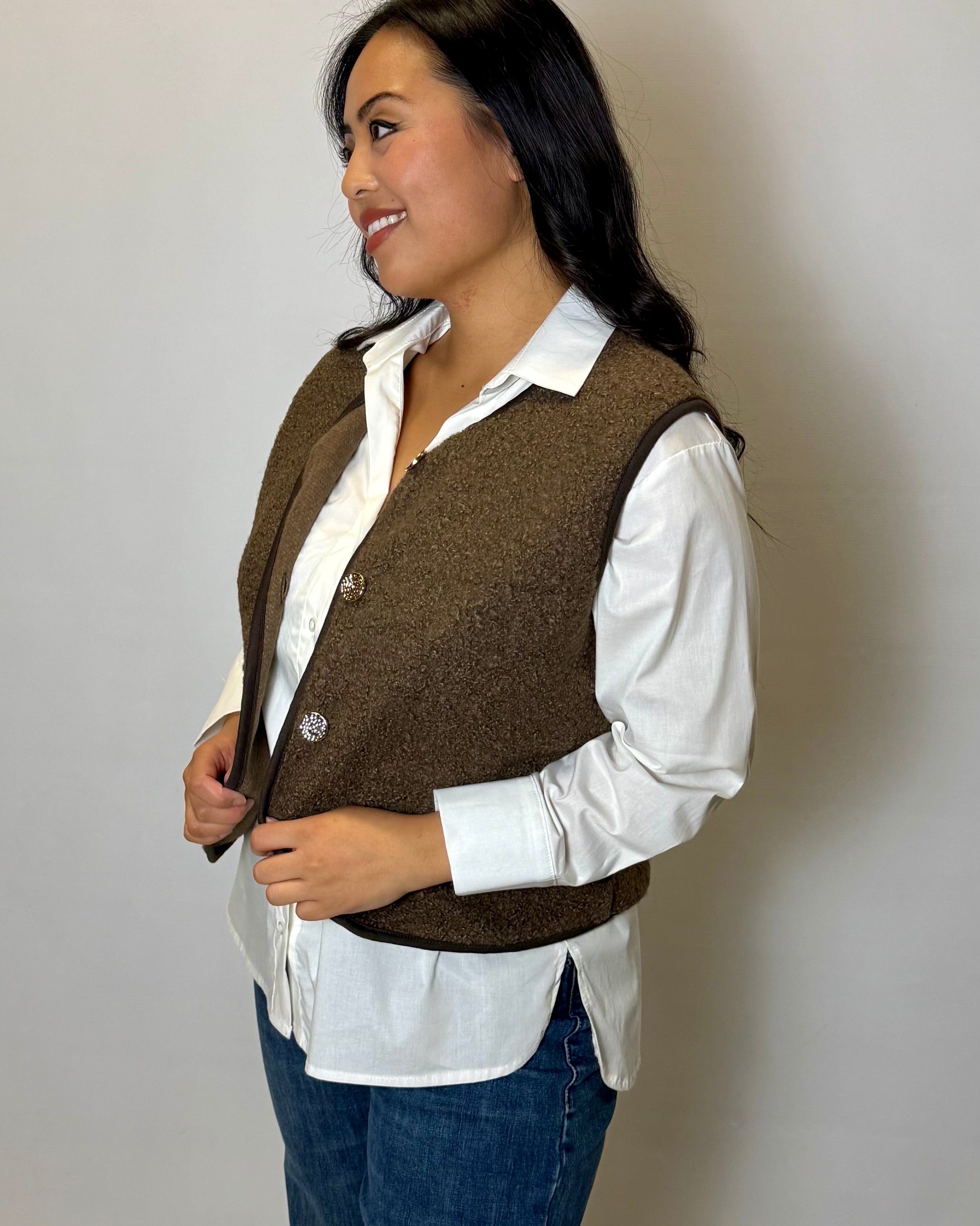 Woman wearing a brown vest over a white shirt and blue jeans against a plain background