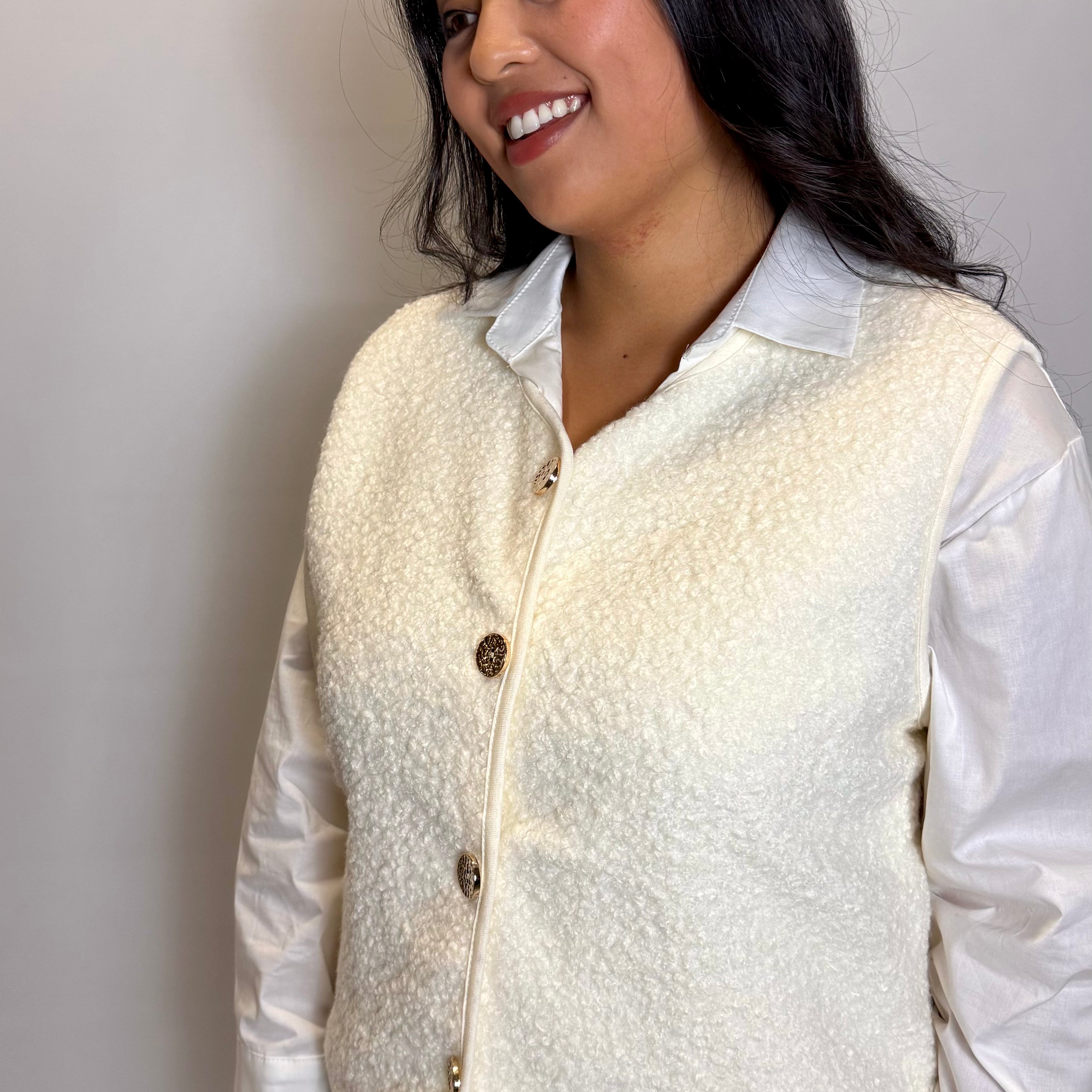 Woman wearing a cream-colored vest over a white shirt against a plain background