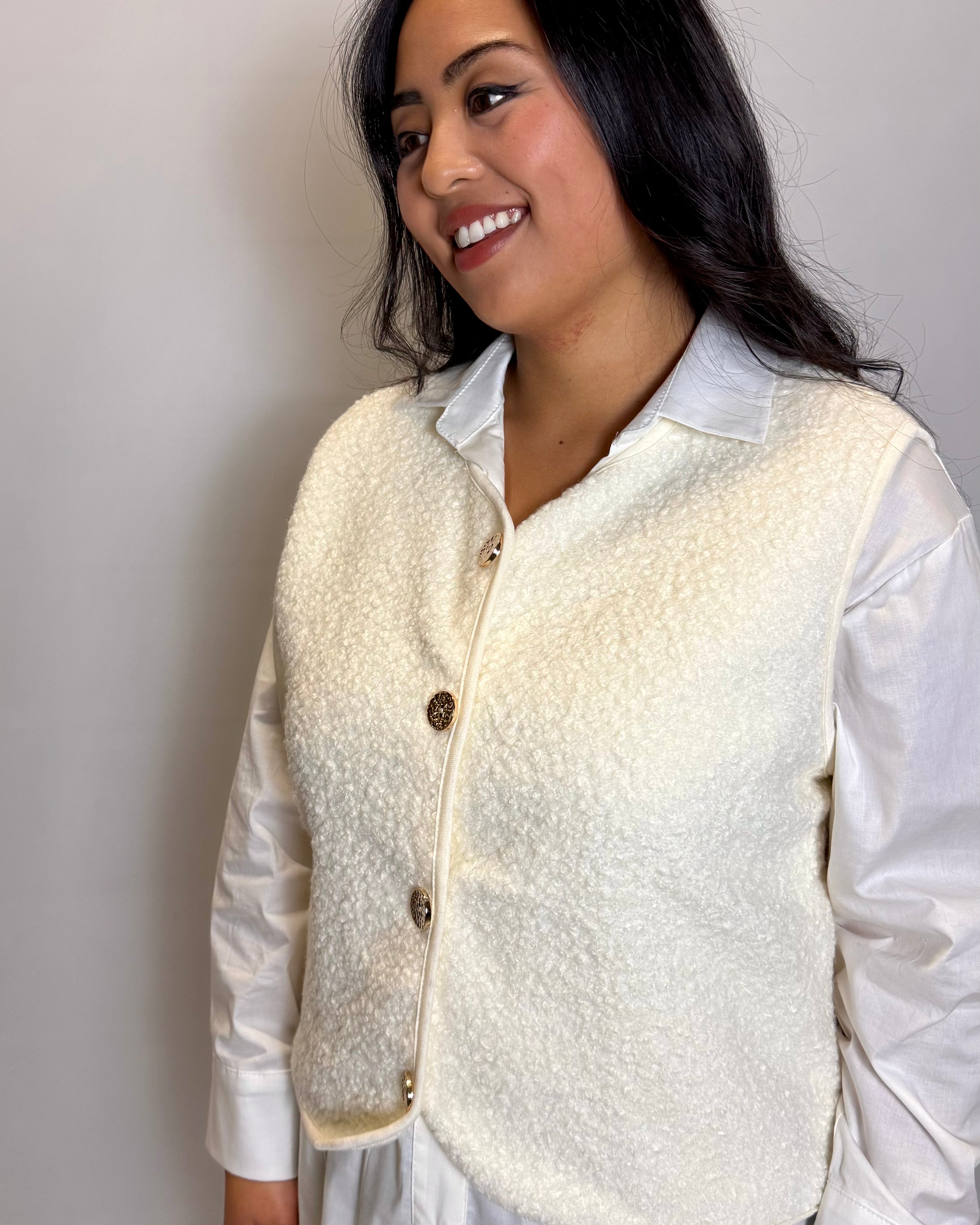 Woman wearing a cream-colored vest over a white shirt against a plain background