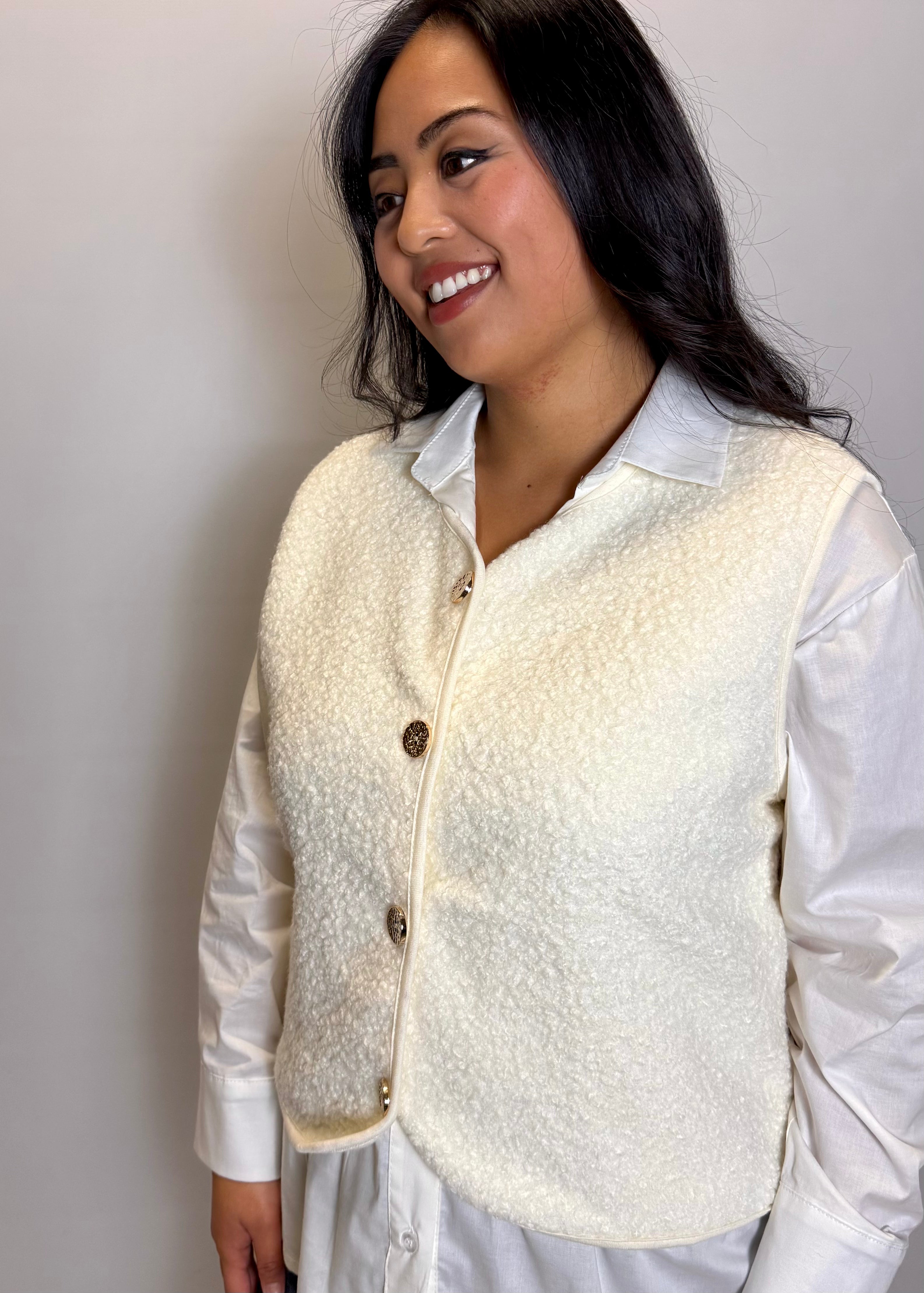 Woman wearing a cream-colored vest over a white shirt against a plain background
