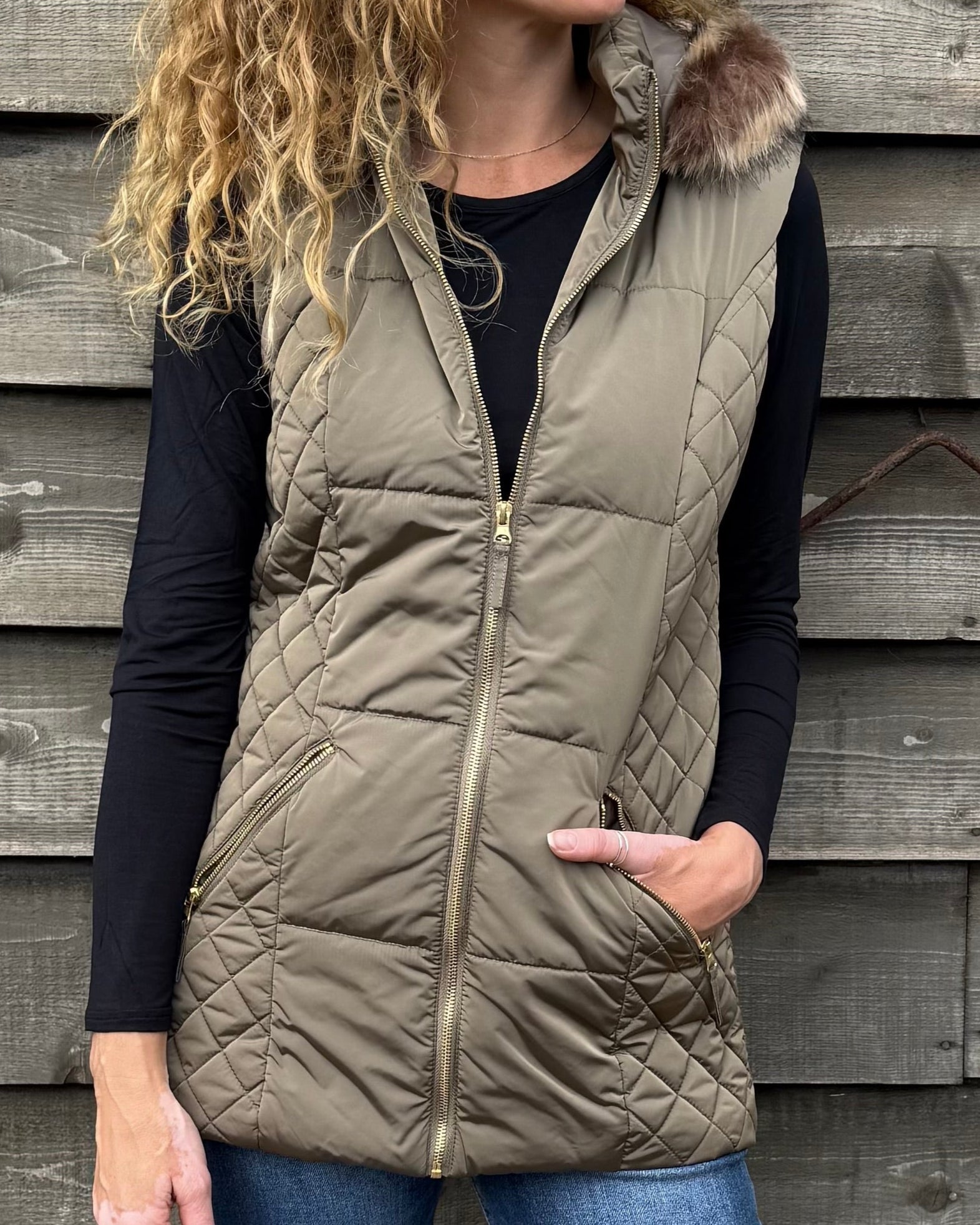 Woman wearing a beige puffer vest standing against a wooden wall.