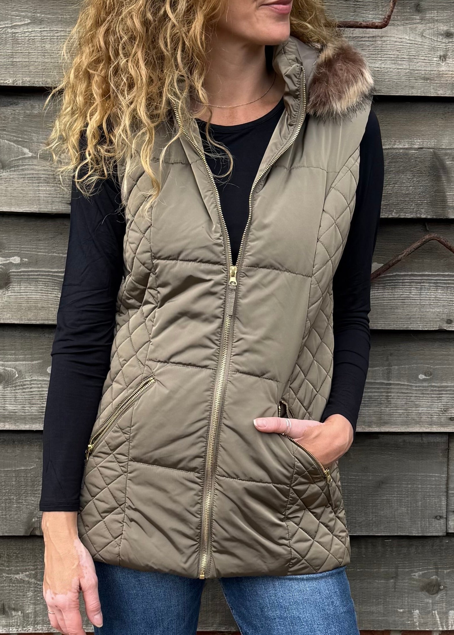 Woman wearing a beige puffer vest standing against a wooden wall.