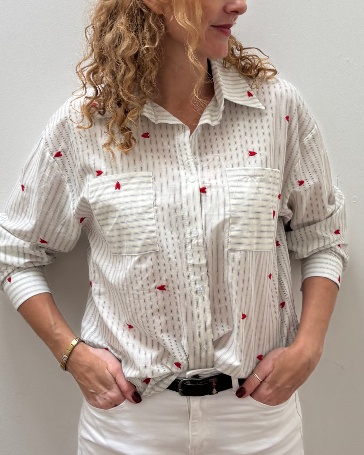 A woman with curly blonde hair stands against a plain light gray wall wearing a white long-sleeve button-up shirt with thin vertical gray stripes and small red embroidered motifs, paired with white pants and a black belt.