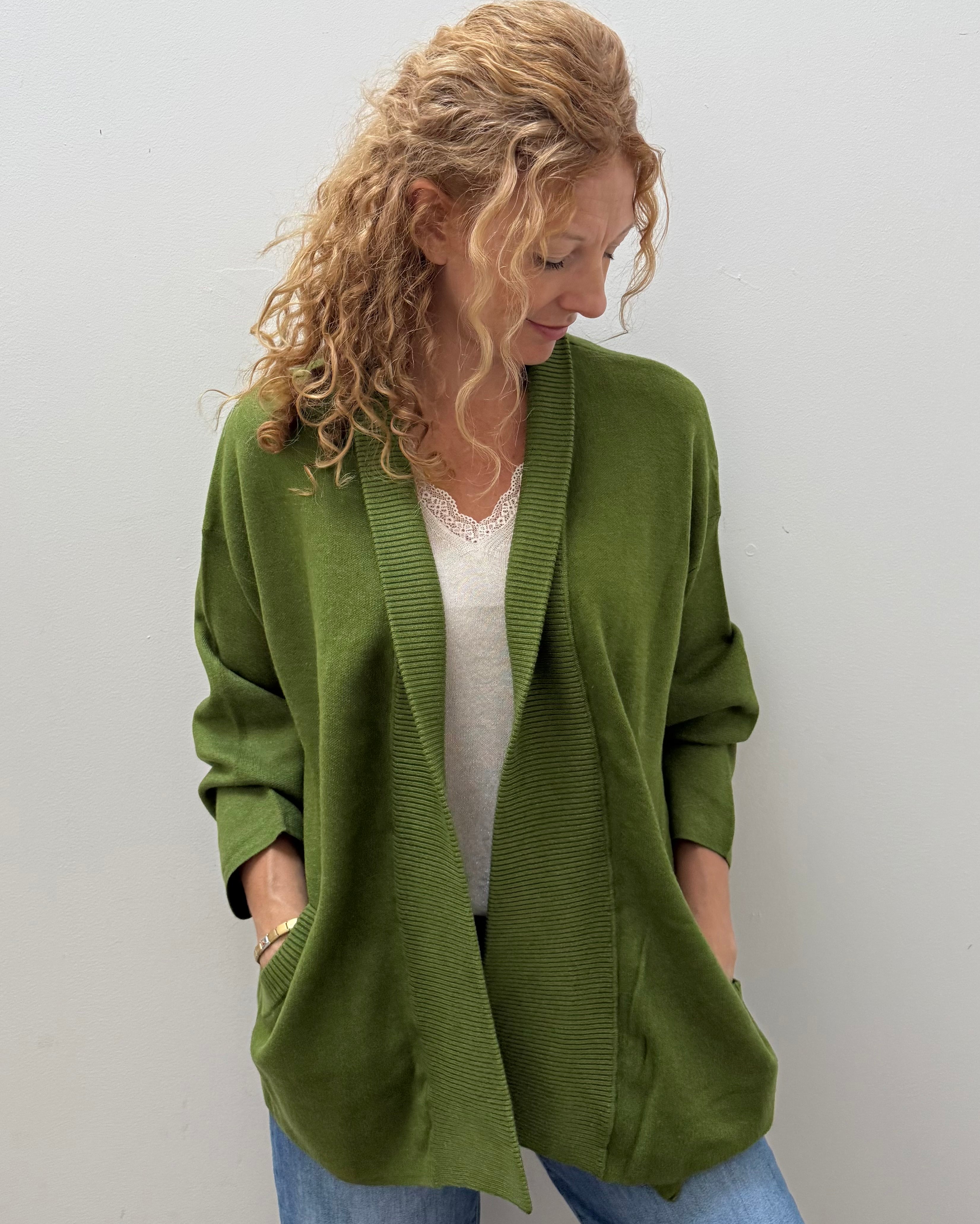 A woman with curly blonde hair wearing an olive green open-front cardigan over a white top and blue jeans, standing against a plain light gray wall.