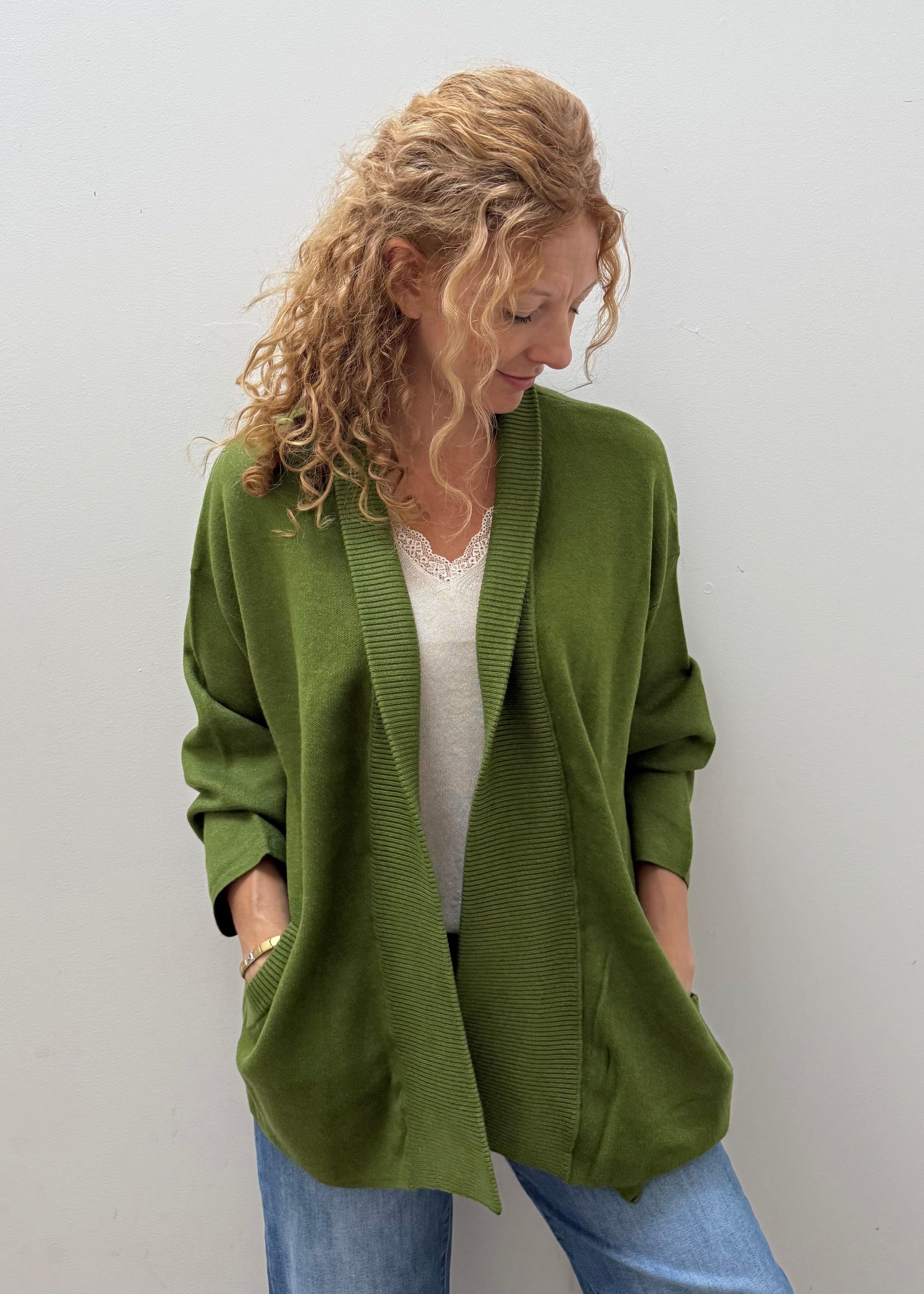 A woman with curly blonde hair wearing an olive green open-front cardigan over a white top and blue jeans, standing against a plain light gray wall.