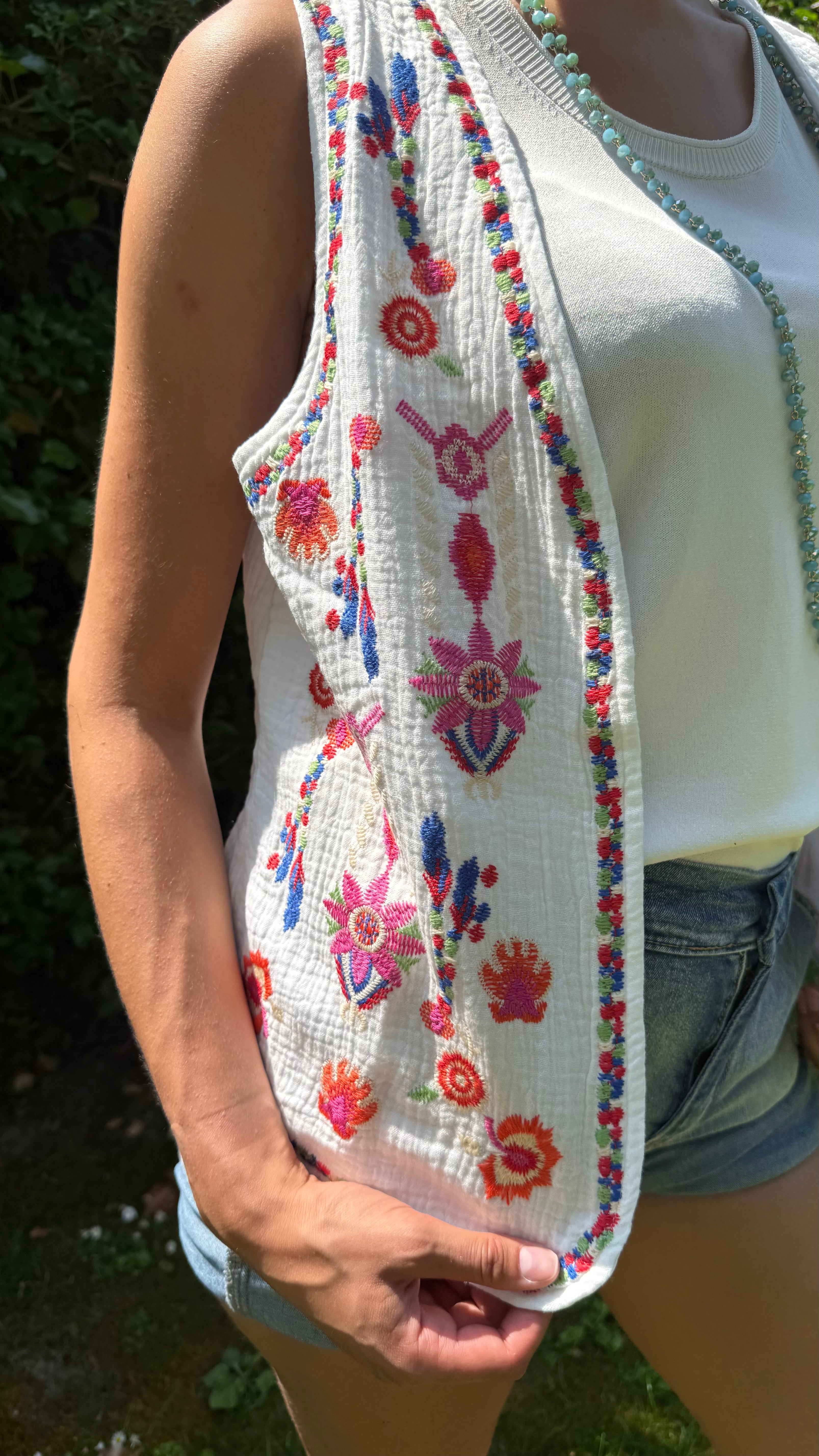Person wearing a floral embroidered vest over a white tank top with denim shorts.