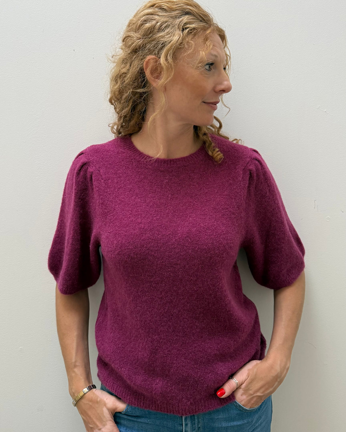 A woman with blonde curly hair wearing a short-sleeved, textured magenta knit sweater and blue jeans, standing against a plain light gray wall.