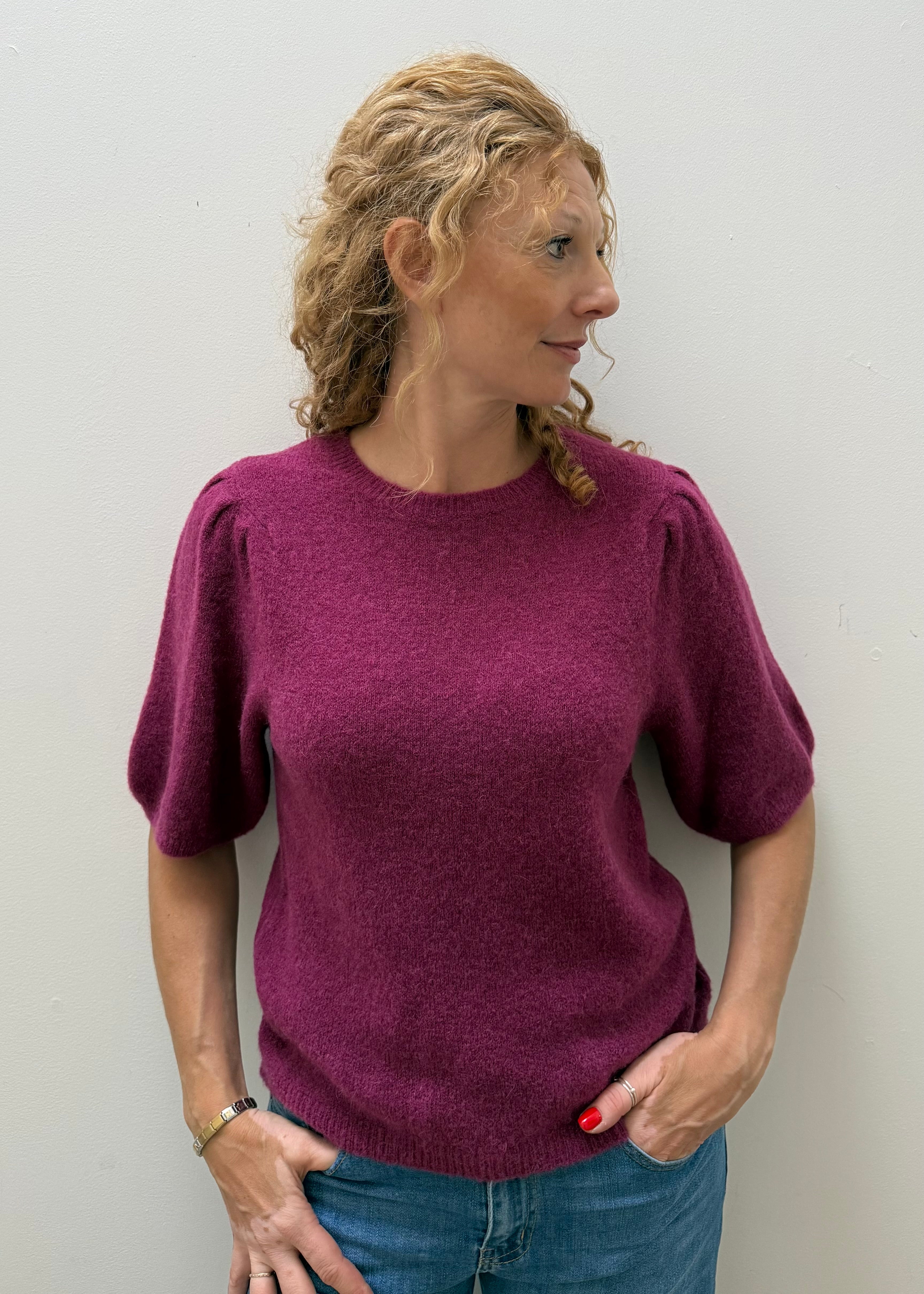 A woman with blonde curly hair wearing a short-sleeved, textured magenta knit sweater and blue jeans, standing against a plain light gray wall.