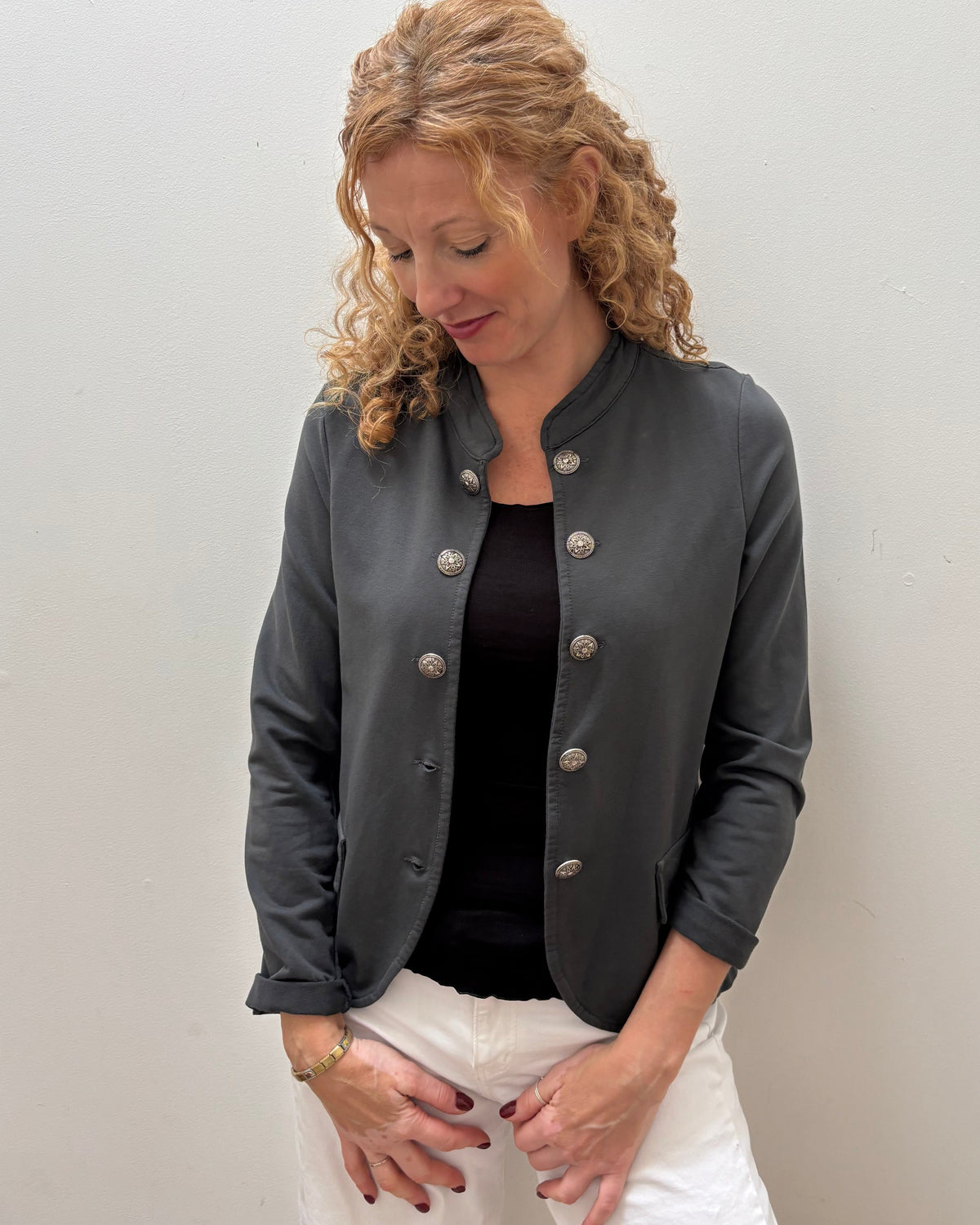 A woman with curly blonde hair wearing a dark gray leather jacket with silver buttons, a black top, and white pants, standing against a plain light gray wall.